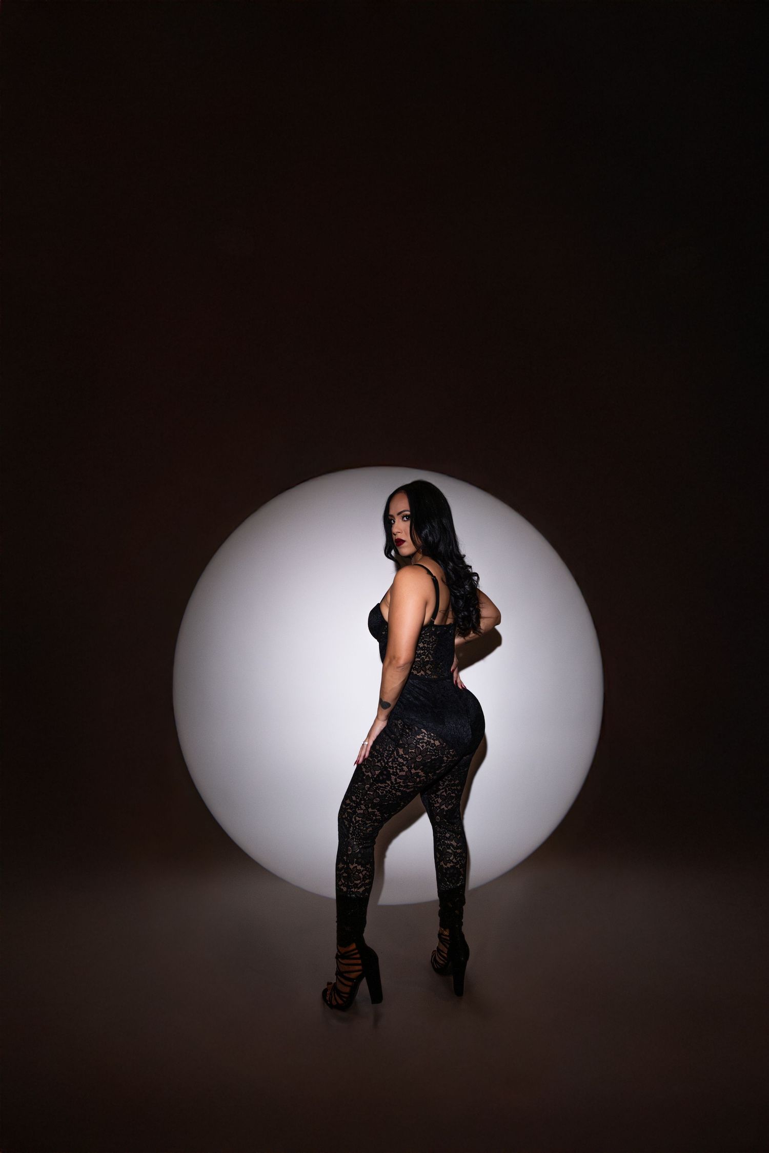 Woman in black lace bodysuit poses confidently in front of glowing circular light on dark background.