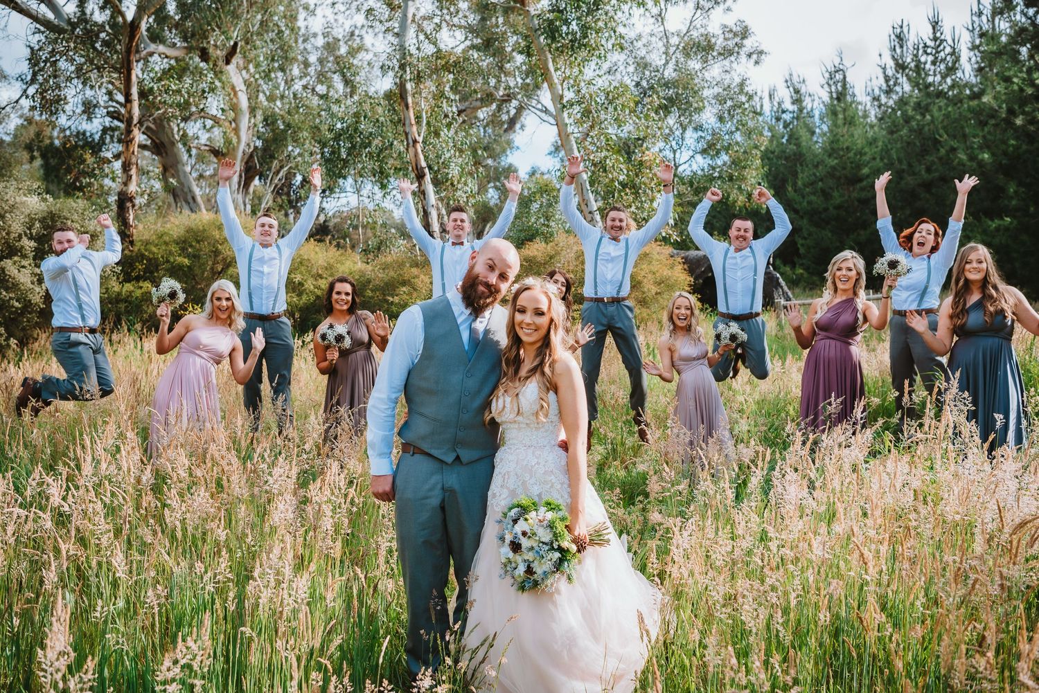 A wedding party celebrates in a sun-drenched meadow with tall grass and eucalyptus trees in the background.