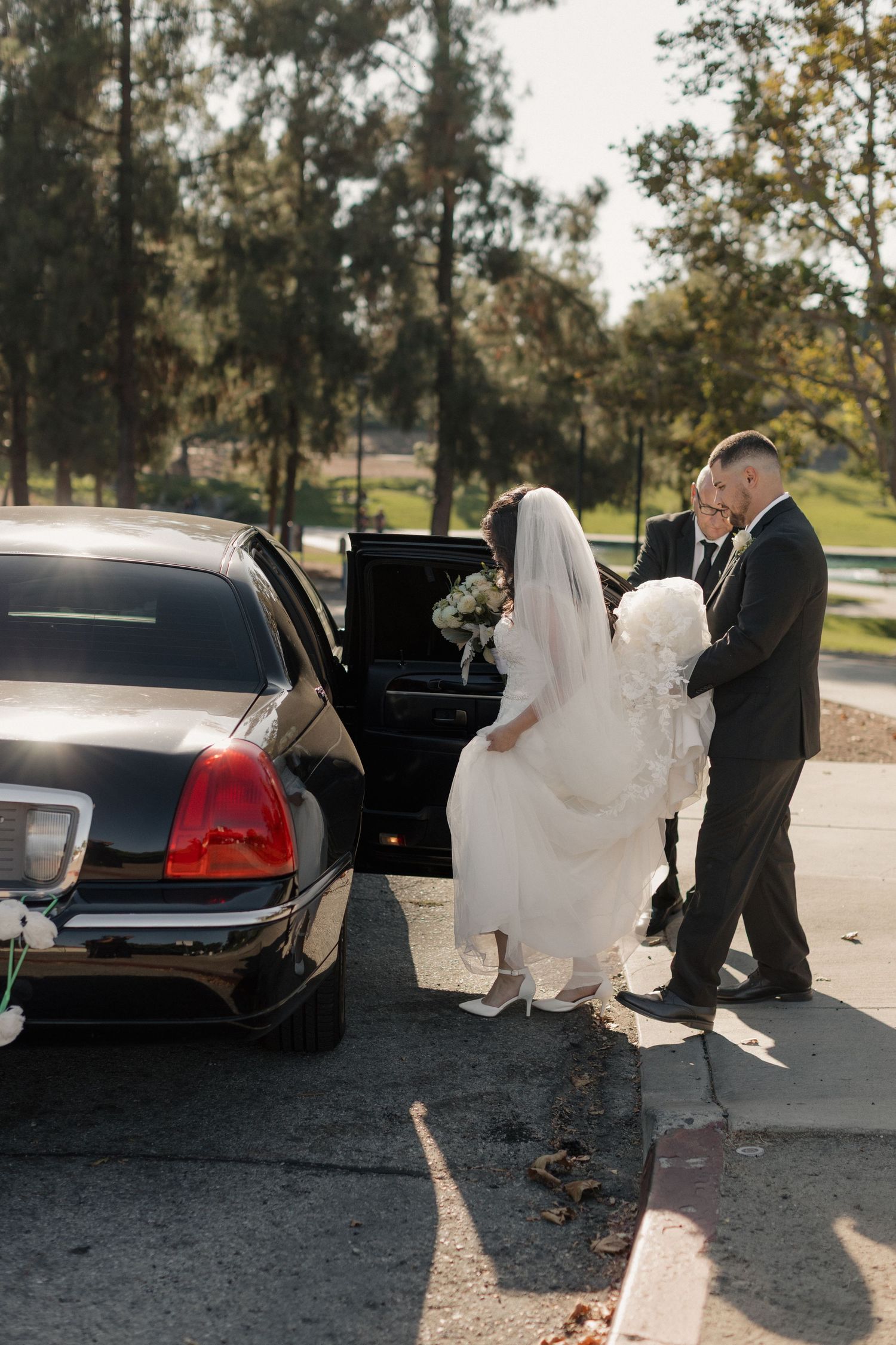 Newlyweds near a black car outdoors with sunlight creating dramatic shadows.