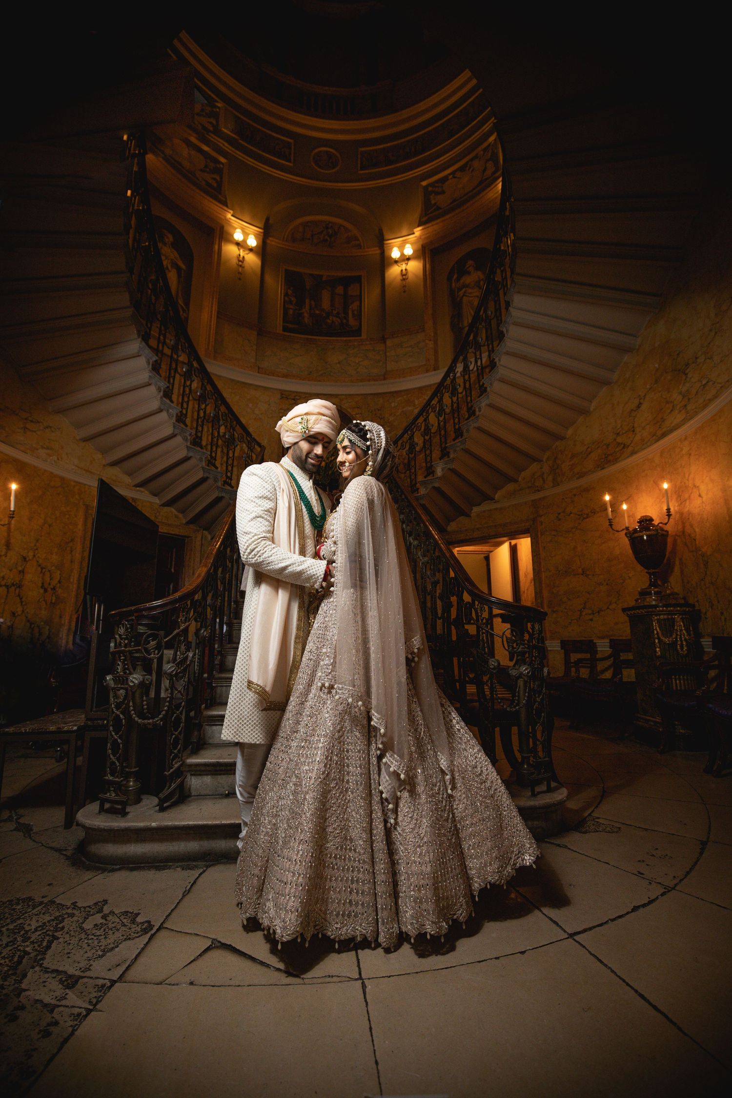 Romantic wedding couple standing in front of an elegant curved staircase lit by warm sconces in an ornate mansion.