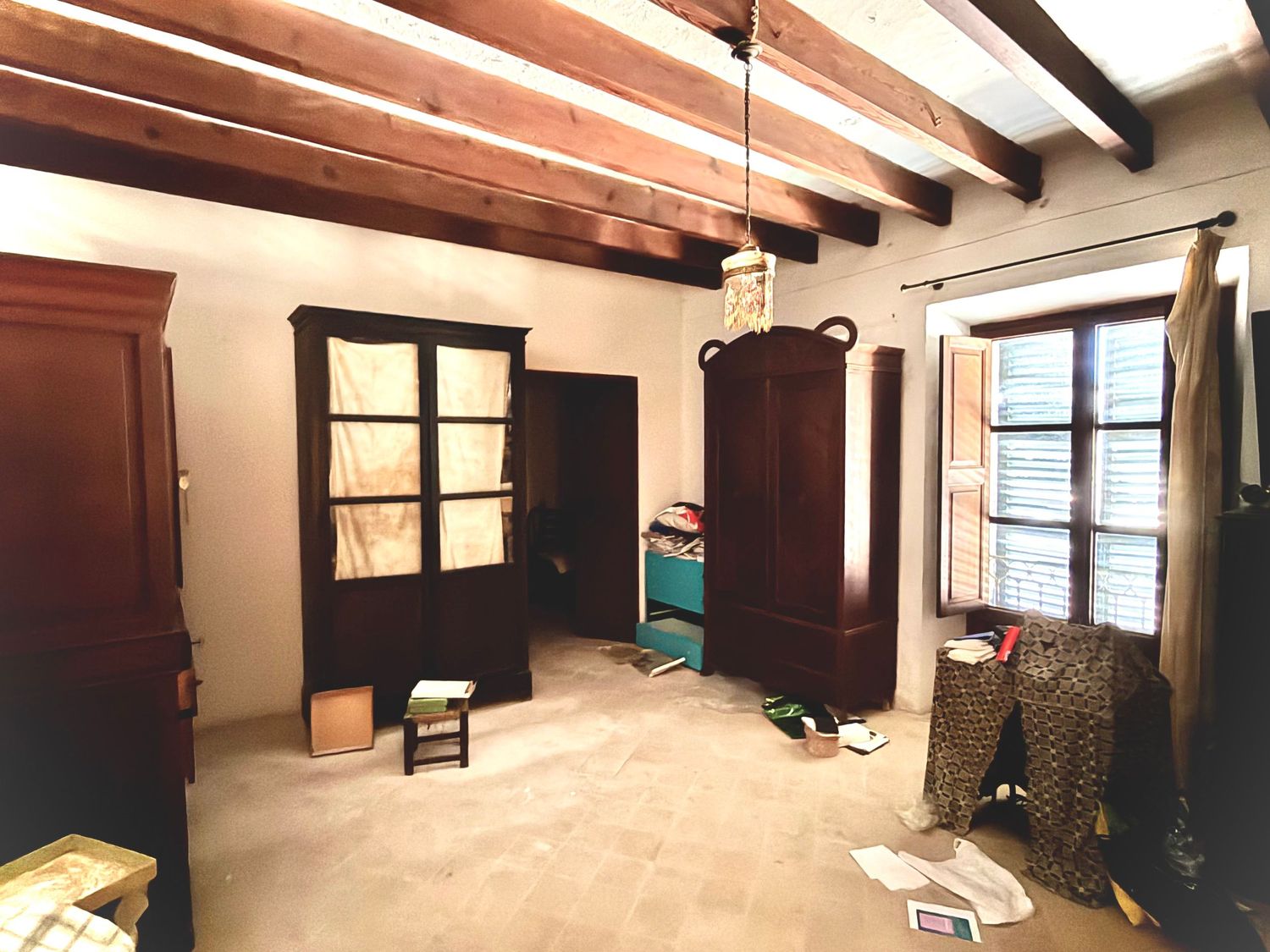 Rustic room with wooden beam ceiling, dark wood armoire, tiled floor, and natural light from shuttered windows.