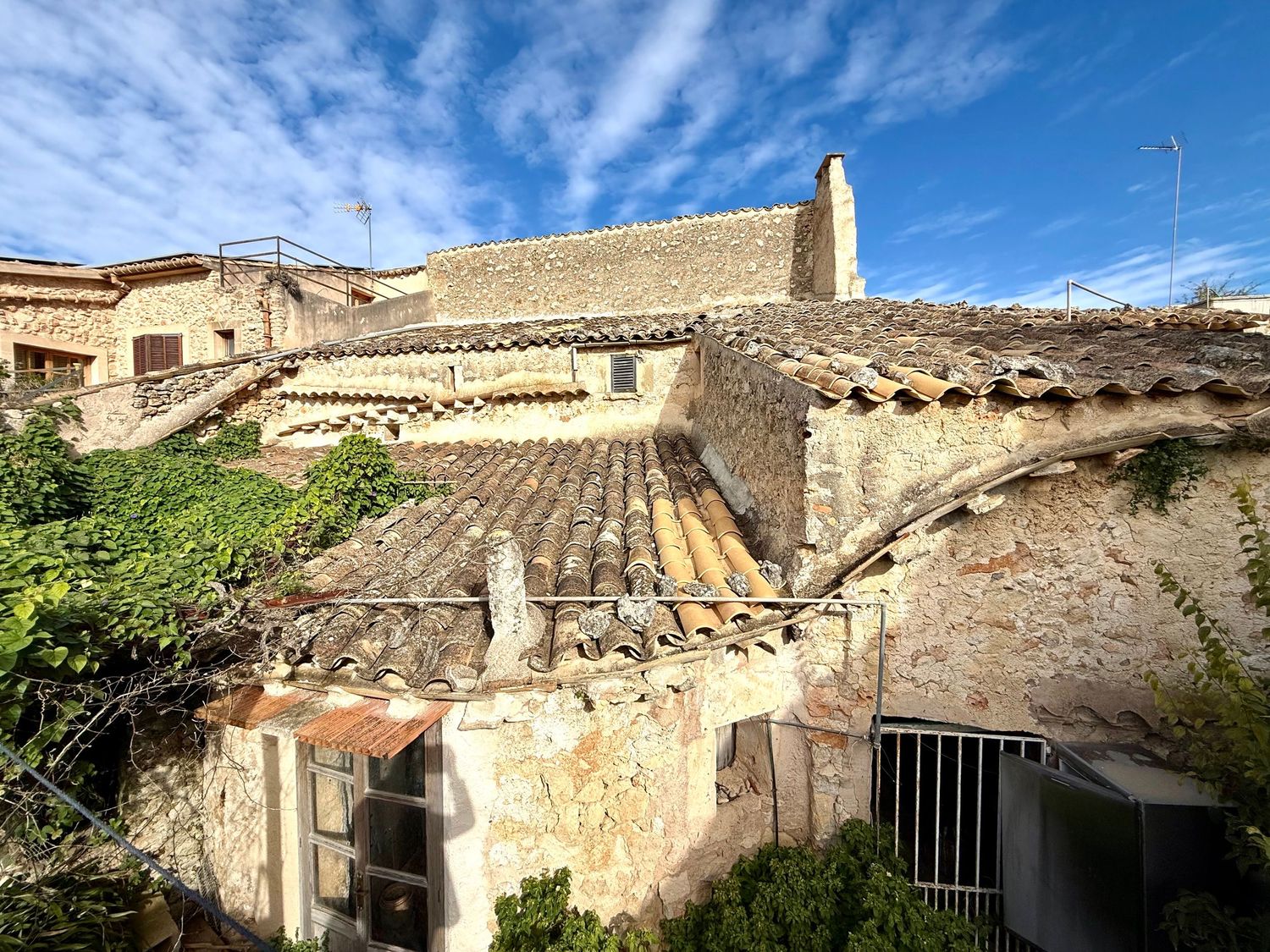 Stone building with terracotta roof tiles under blue sky, surrounded by lush green ivy and vegetation in a rustic Mediterranean village.