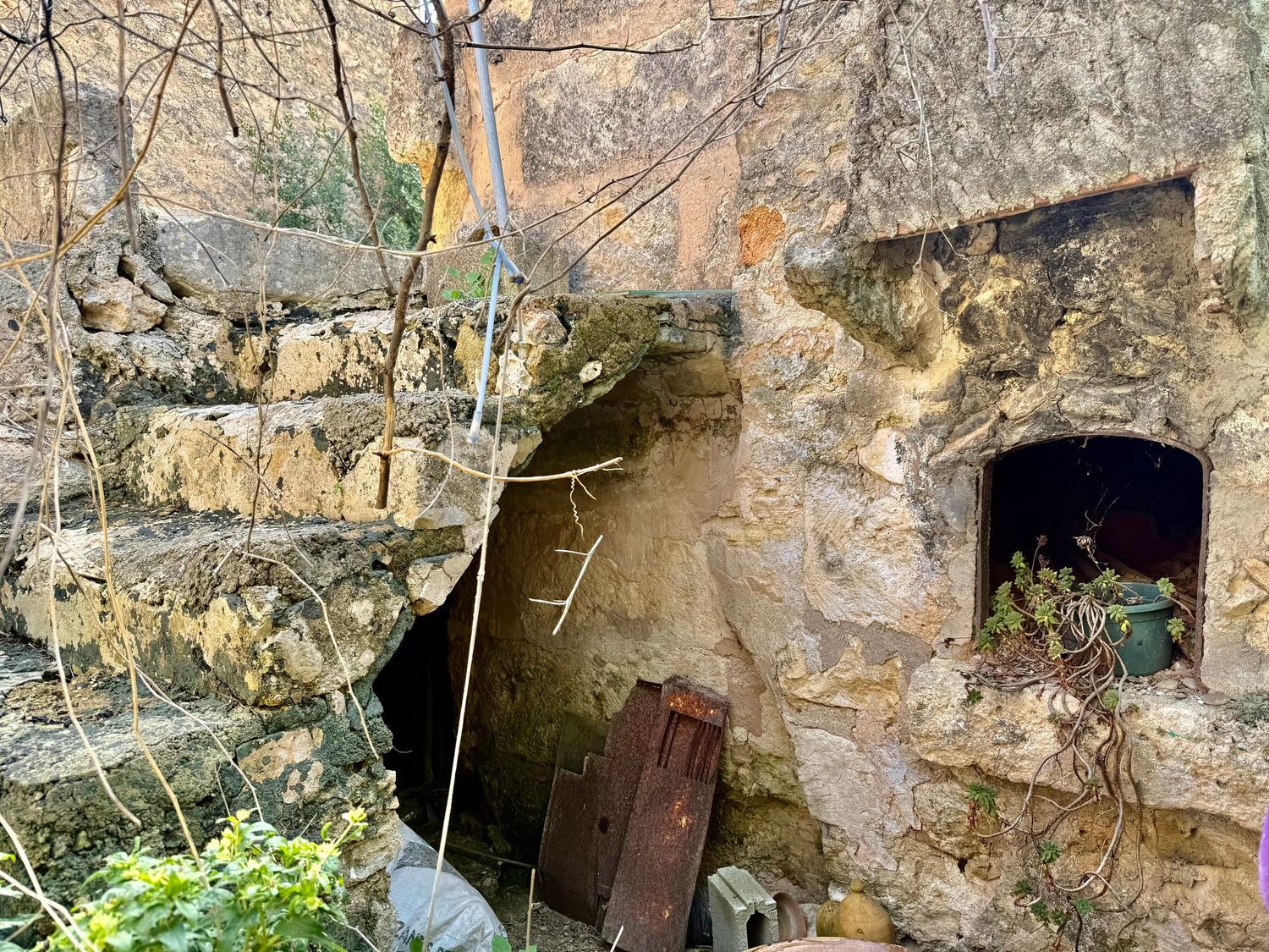 Ancient stone ruins with weathered walls, wooden door, and overgrown vegetation in a historic Mediterranean setting.
