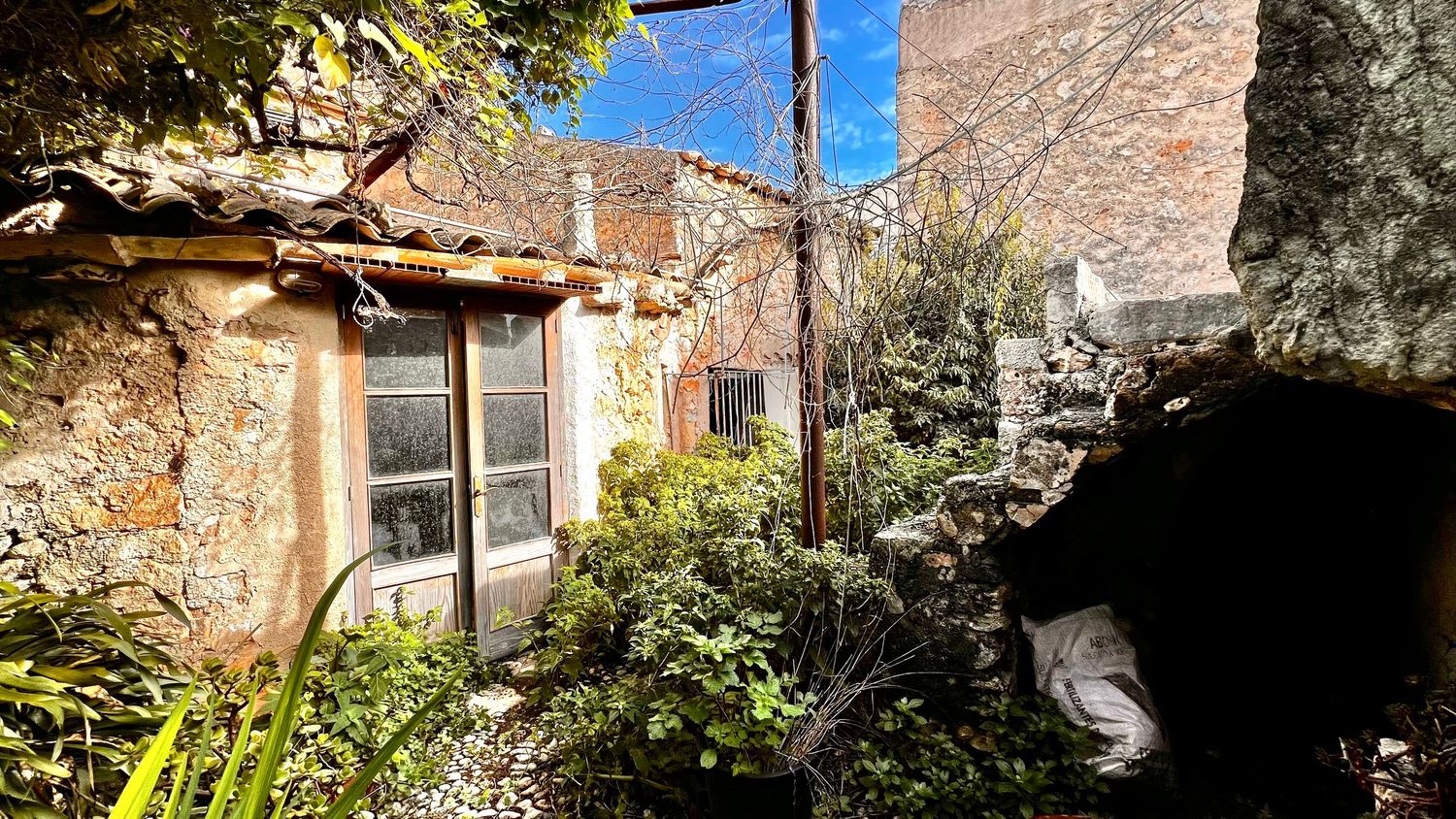 Rustic stone house exterior with lush garden, weathered walls, and vintage window under a clear blue sky.