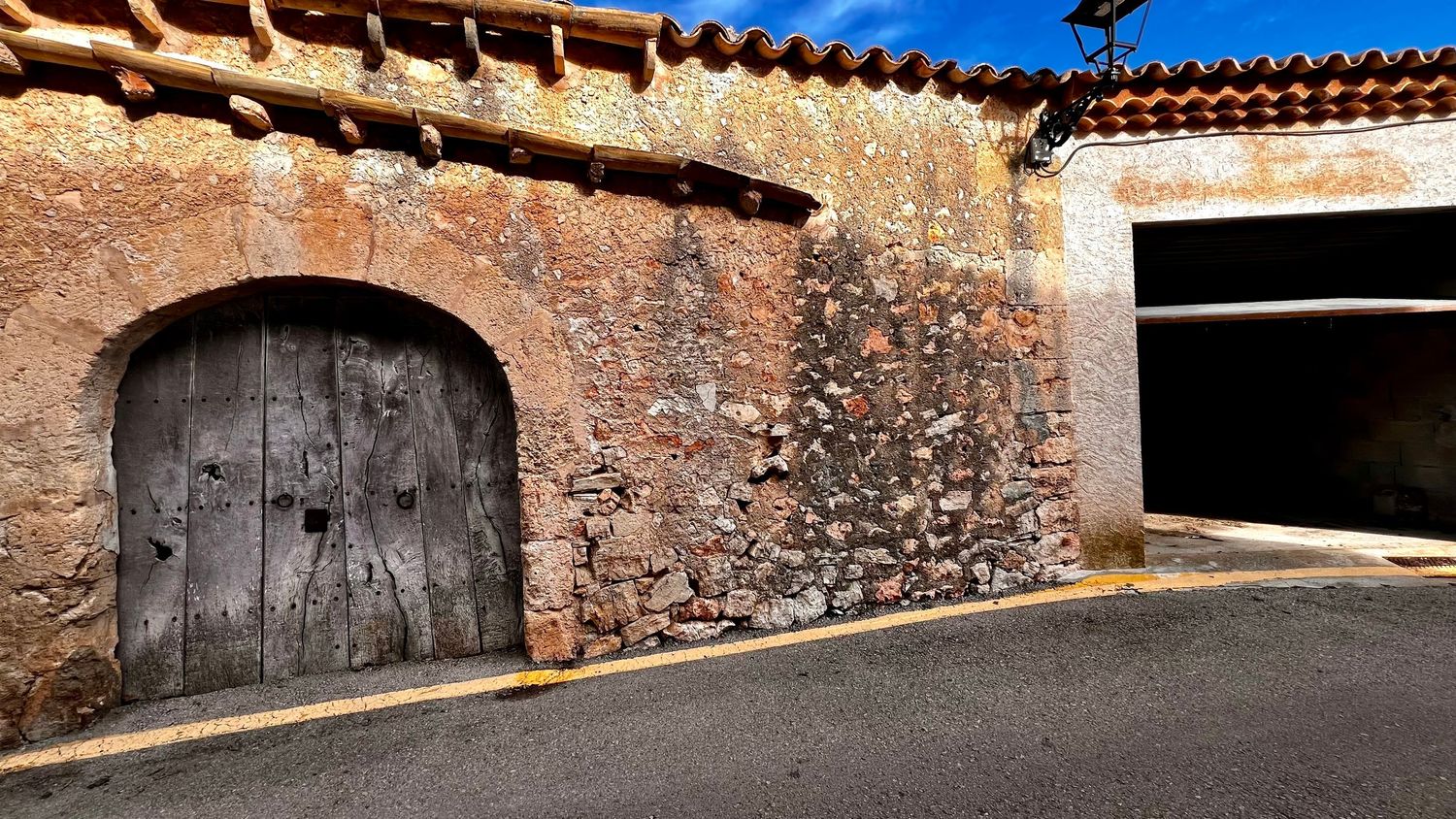 Ancient stone wall with weathered wooden arched door beside a modern garage entrance in a rustic Mediterranean village.