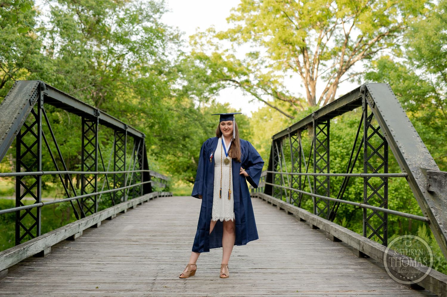 Graduate in academic regalia poses on rustic bridge in park setting.