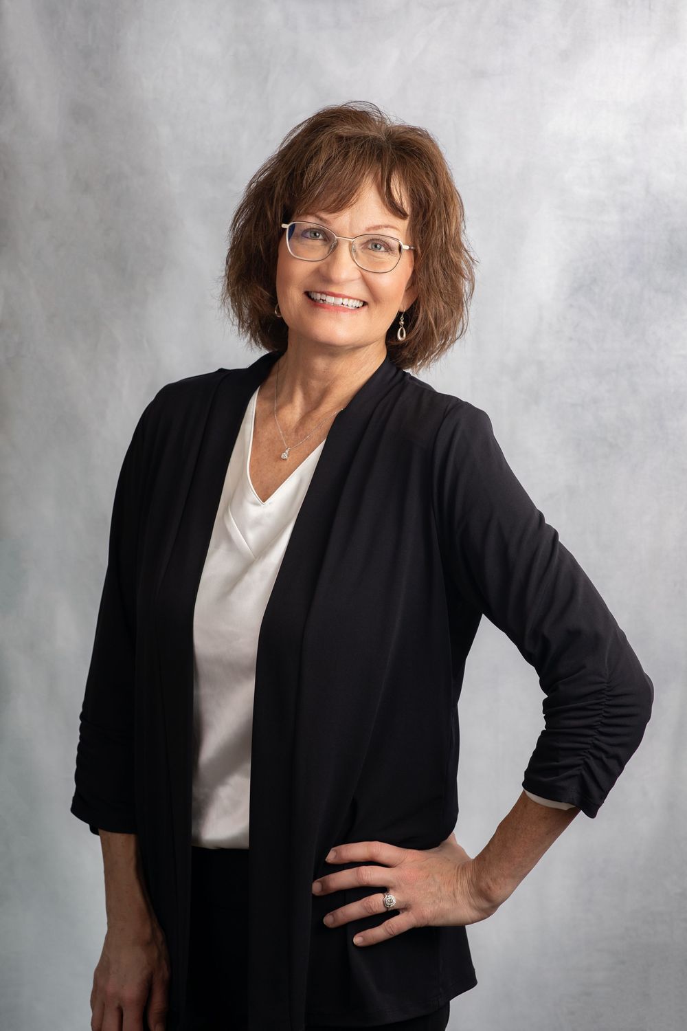 Truly Juli Photography headshot of a female wearing a white shirt and a black cardigan standing in front of a gray backdrop