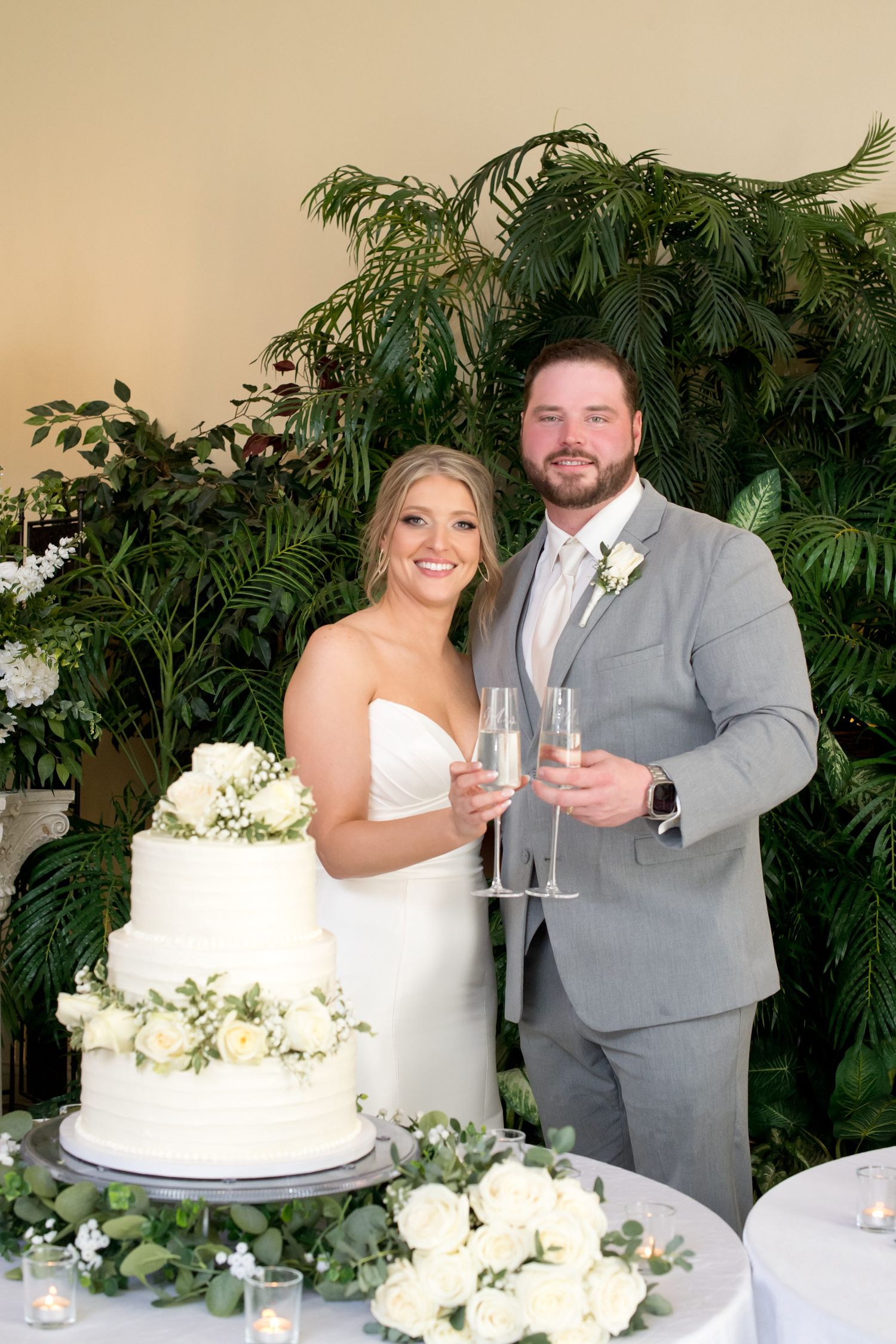 Wedding cake display with white flowers and greenery against tropical plant backdrop.