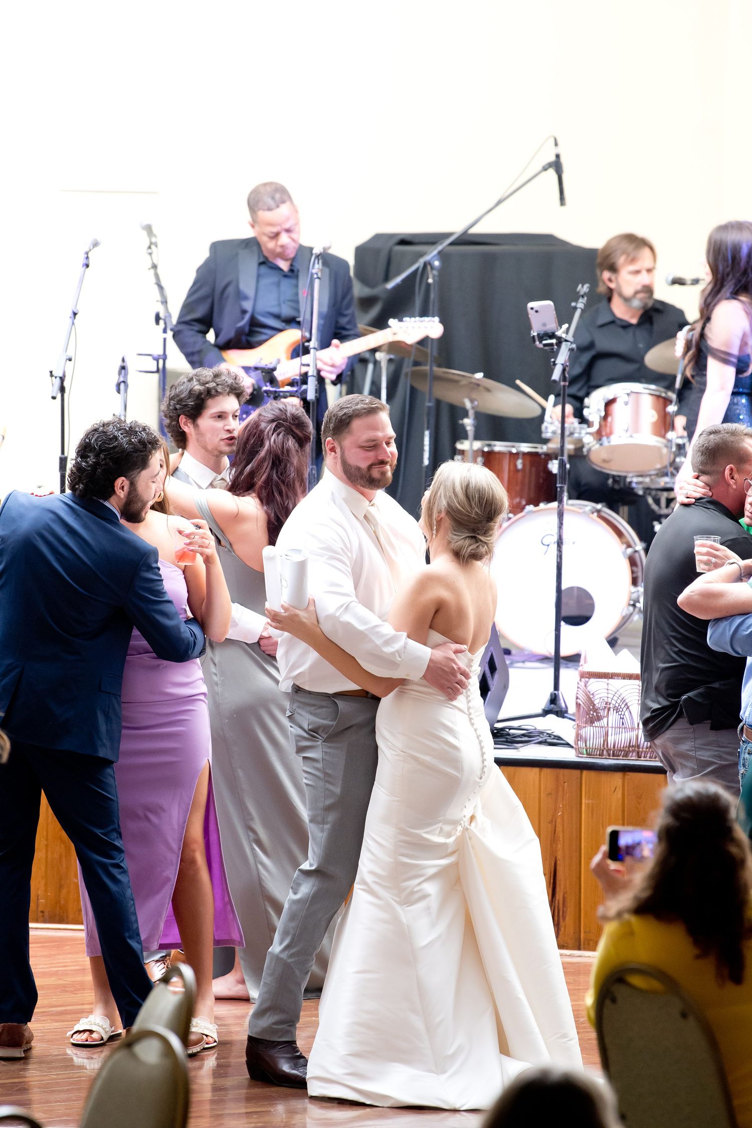Wedding party dancing energetically at reception with live band performing in background.
