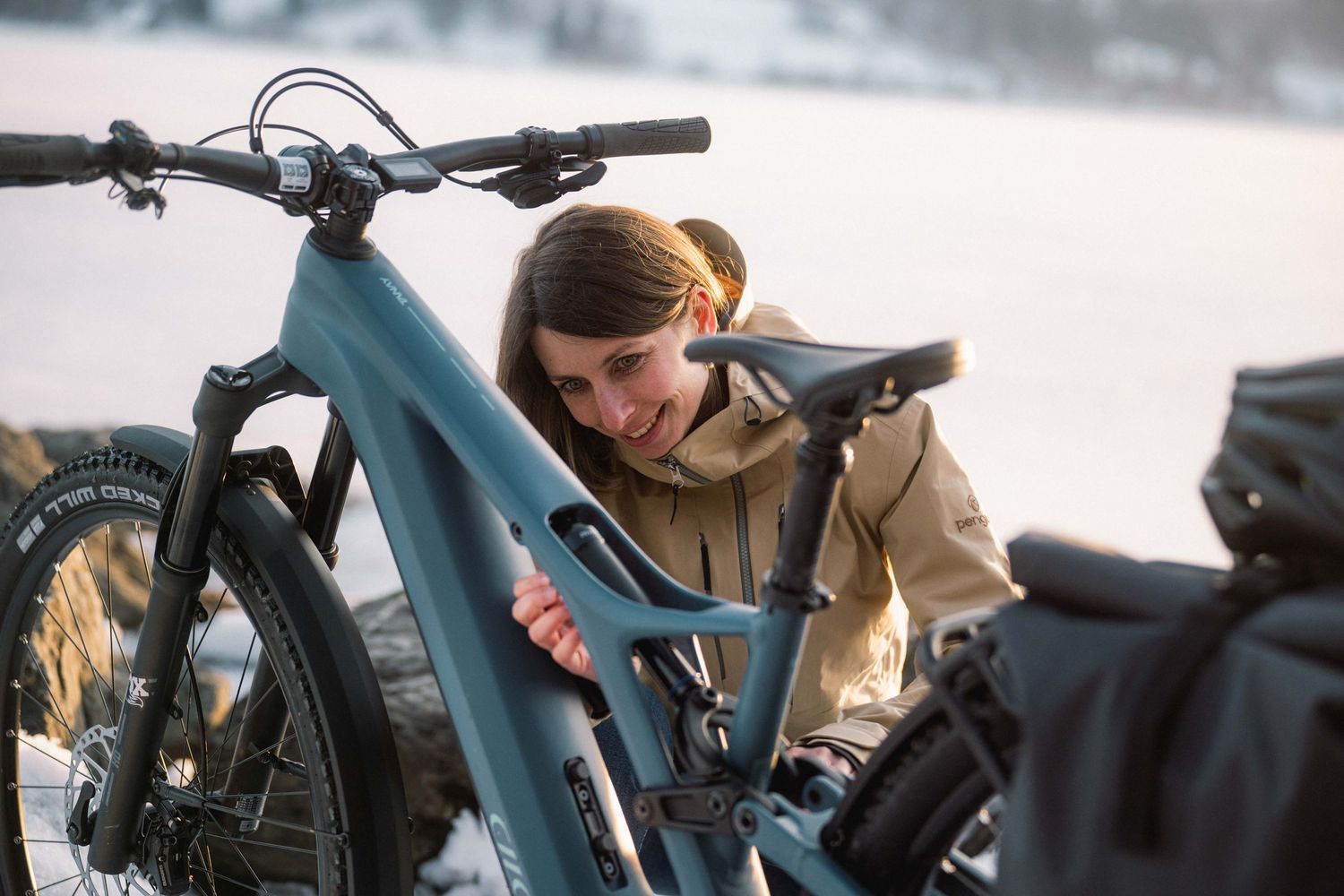 Cyclist in a tan jacket smiles while inspecting a blue mountain bike in snowy winter conditions.