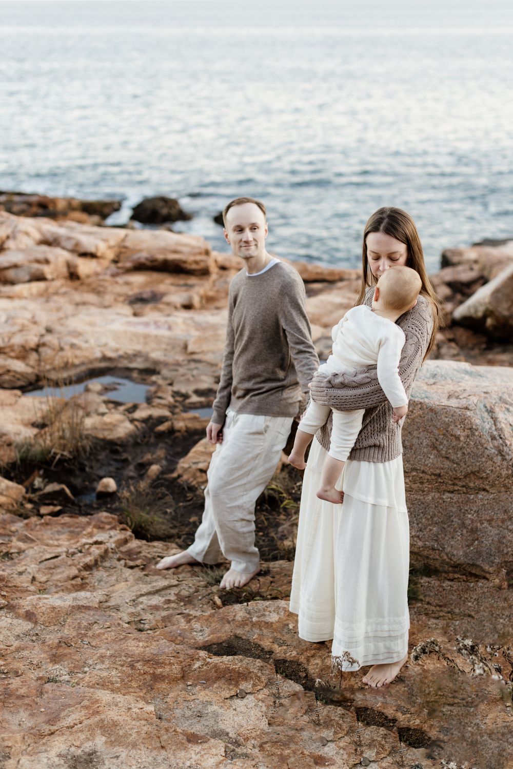 A mother wearing a tan sweater and long cream dress embraces her son as her husband watches on golden rocks in North Shore Gloucester, MA.