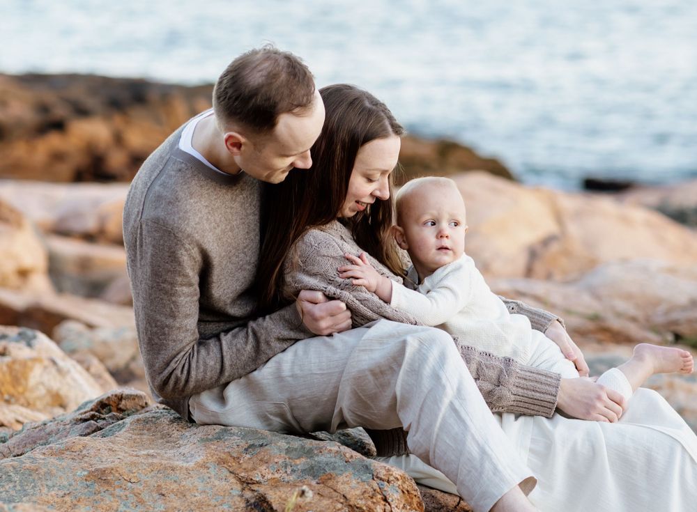 Mother and father wearing tan sweaters gaze at their little boy while lounging on golden  coastal rocks with blue ocean water in background.