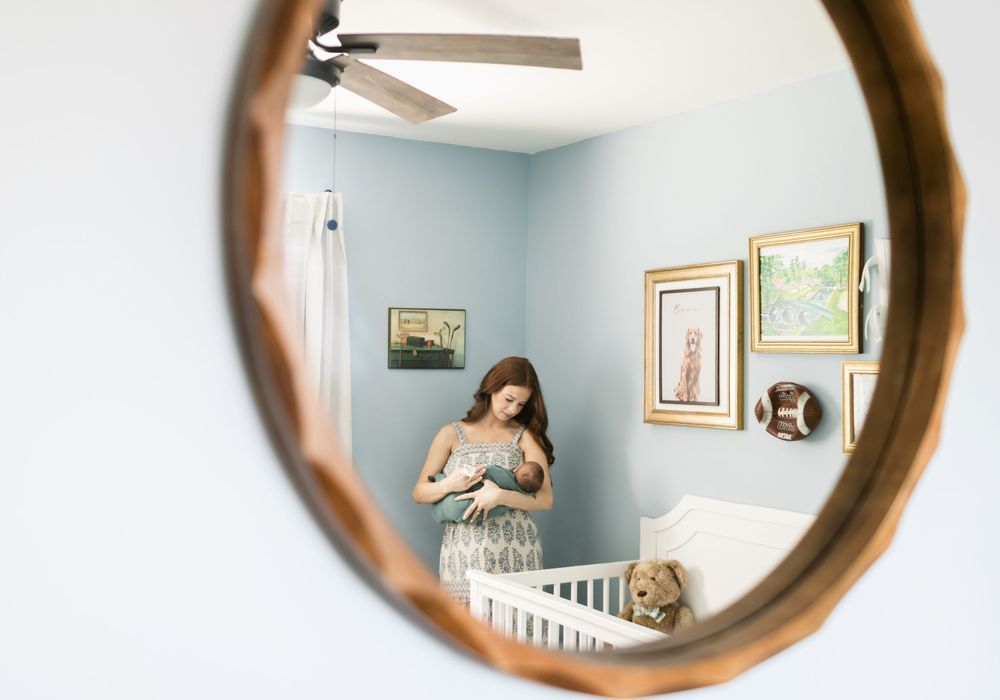 A red-haired mother with long hair holds her newborn son in a mirror reflection in baby's nursery. Monadnock Keene NH newborn photographer.