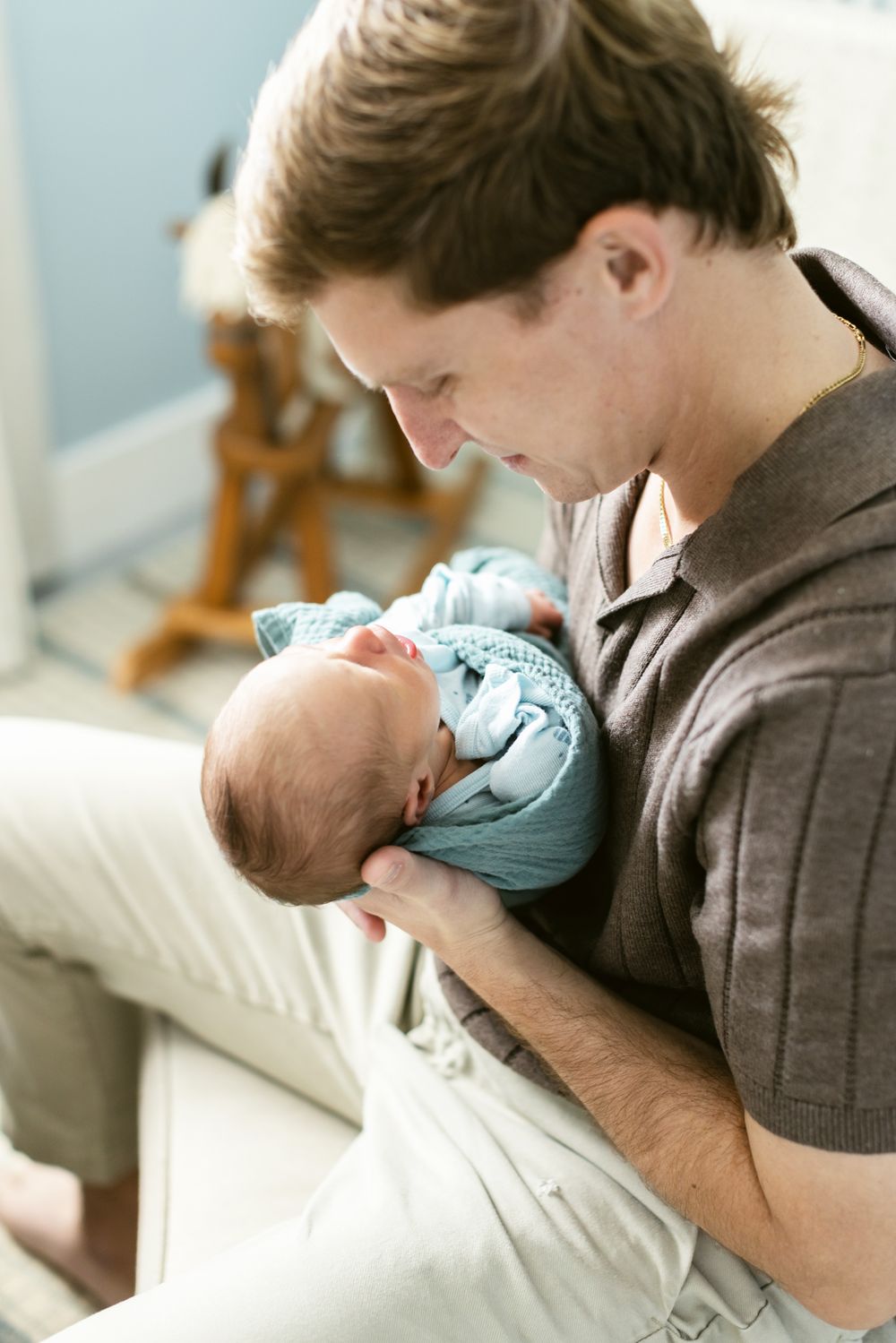 A father wearing a brown short sleeve sweater holds his swaddled newborn for a newborn photo shoot. Concord, Lexington newborn photographer.