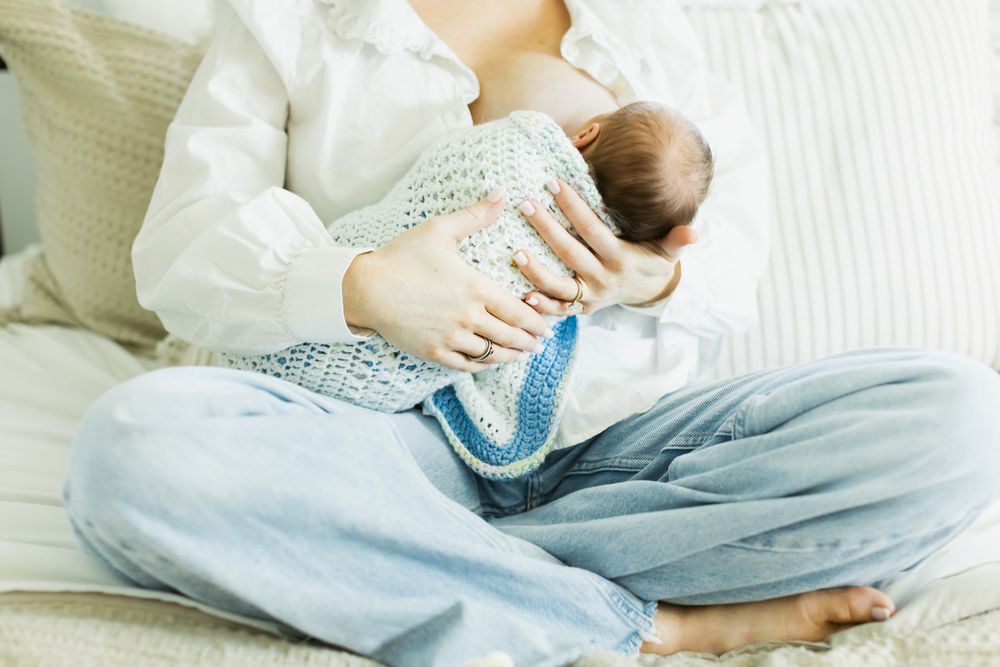 Mother in blue jeans and white peasant shirt breastfeeds newborn swaddled in crochet blanket sitting on bed. Concord Bedford newborn photos.