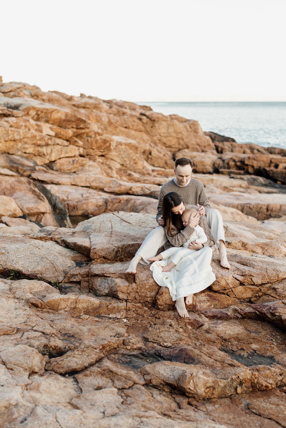 A wife sits between her husband's legs snuggling her baby boy on golden rocks for a family photo shoot in the North Shore, Gloucester MA.
