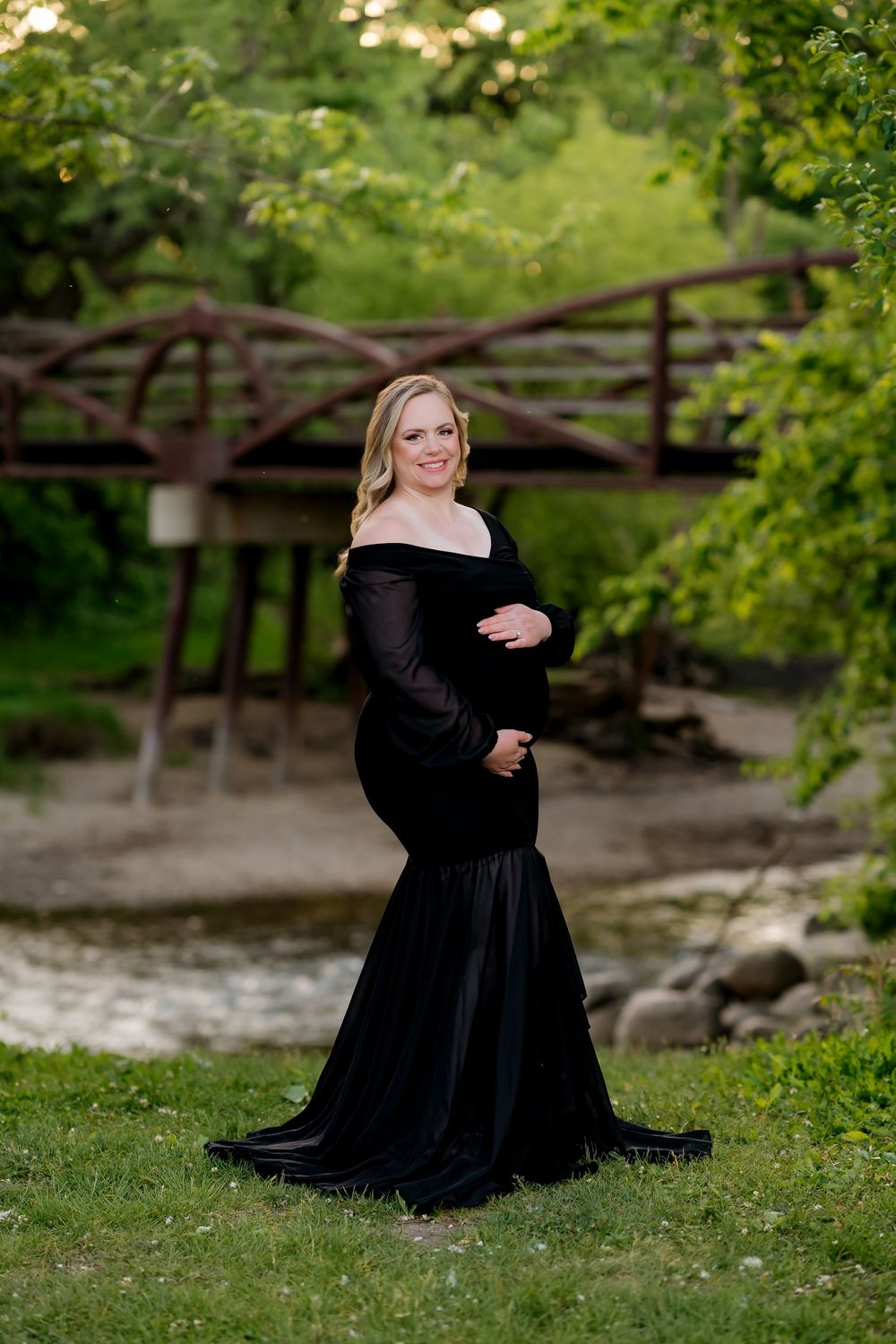 Pregnant woman in elegant black off-shoulder mermaid gown poses outdoors near rustic bridge surrounded by lush green trees.