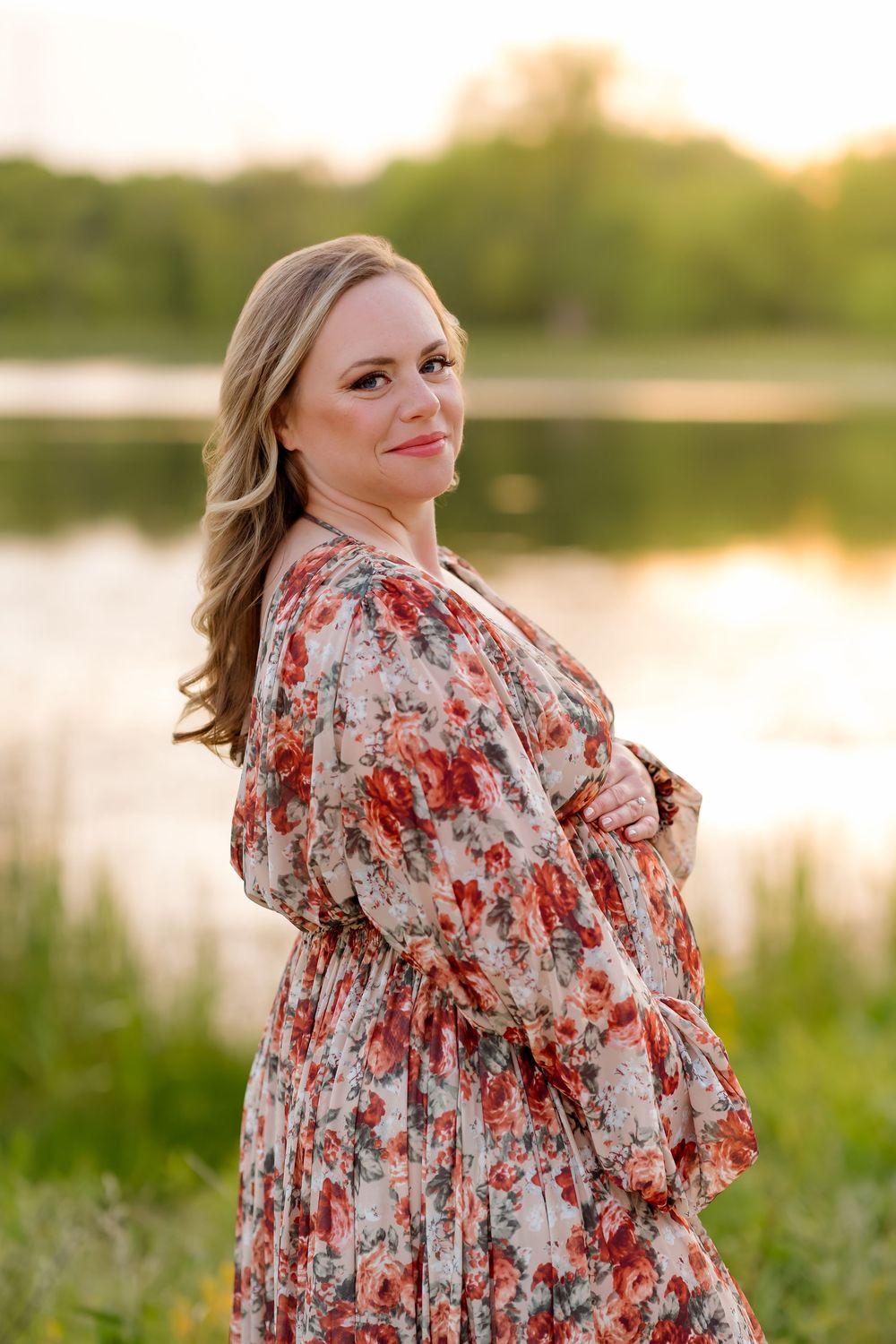 Woman in floral dress posing by a lake at sunset in a lush green outdoor setting.
