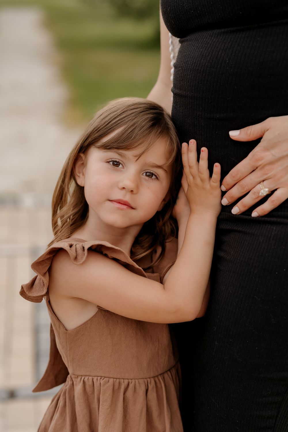 Young girl in brown dress hugs pregnant mother's belly outdoors in a tender maternity photo session.