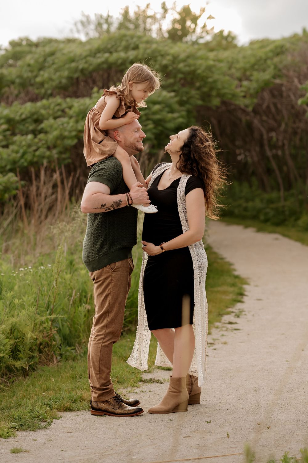 A pregnant woman in a black dress stands with her partner and young daughter on a scenic outdoor path.
