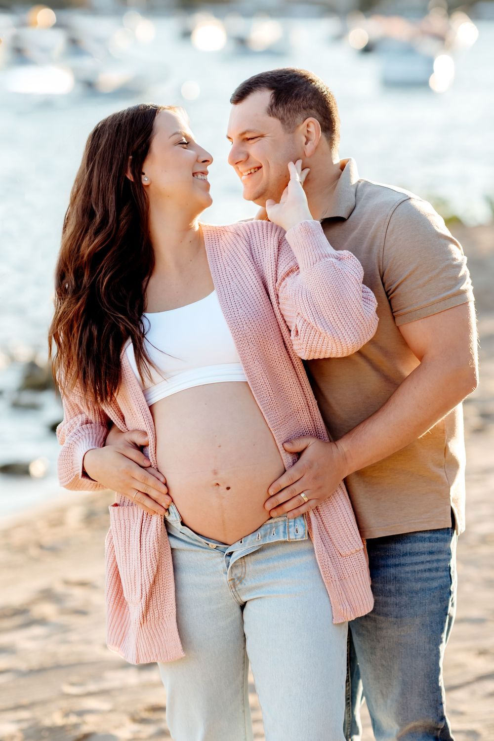 A couple embraces on the beach during a romantic maternity photoshoot in soft natural lighting.