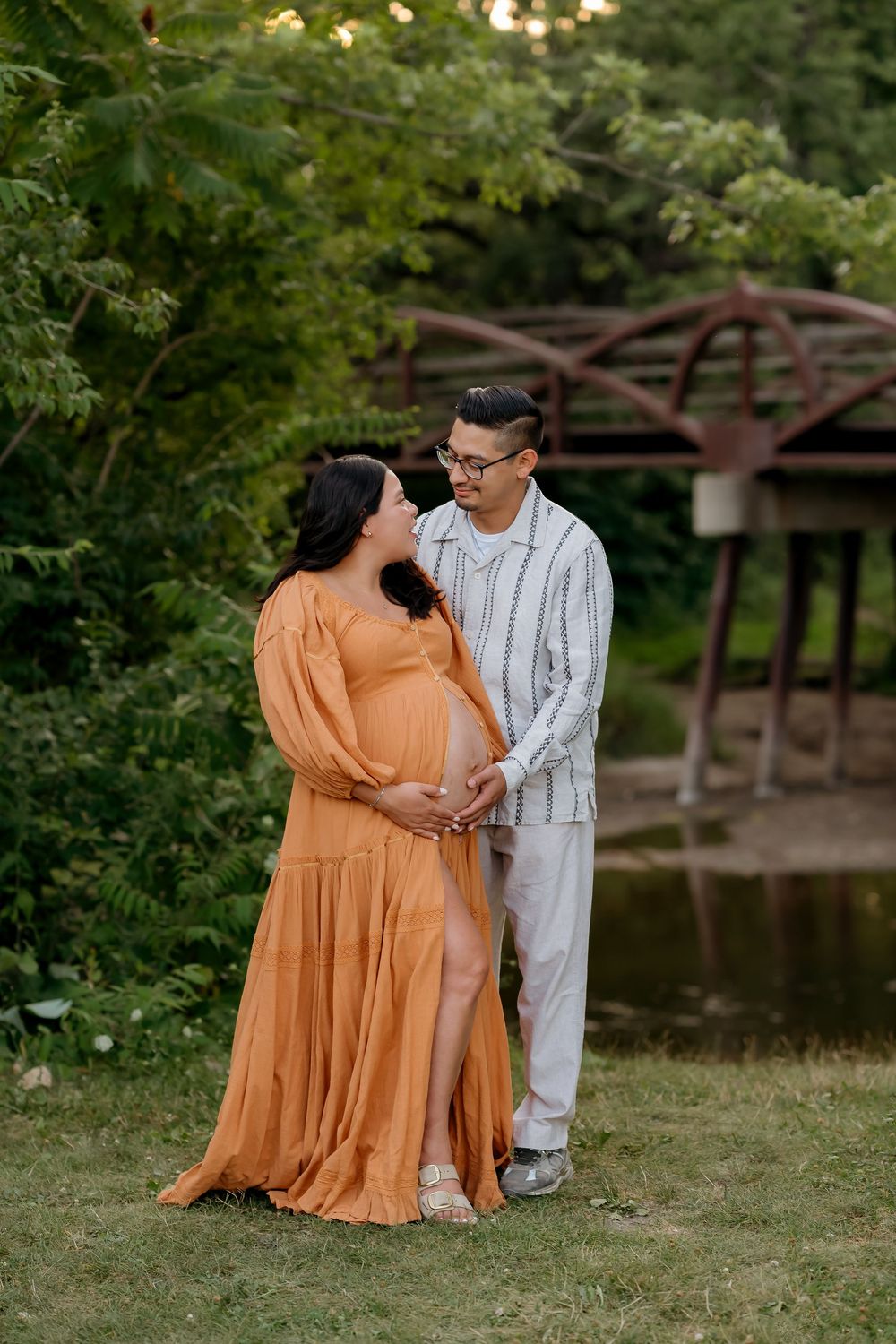 Pregnant couple in orange dress and white shirt holding baby bump near bridge in lush green park setting.