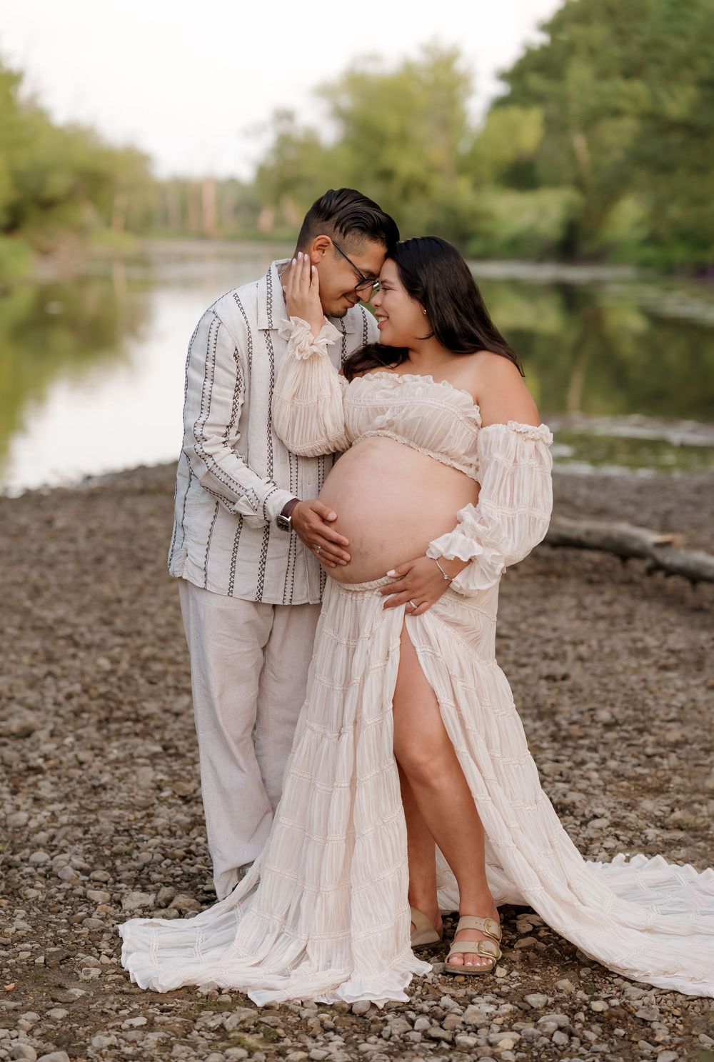 Couple sharing a tender moment during a maternity photo shoot by a river, woman in white lace gown cradling her baby bump.