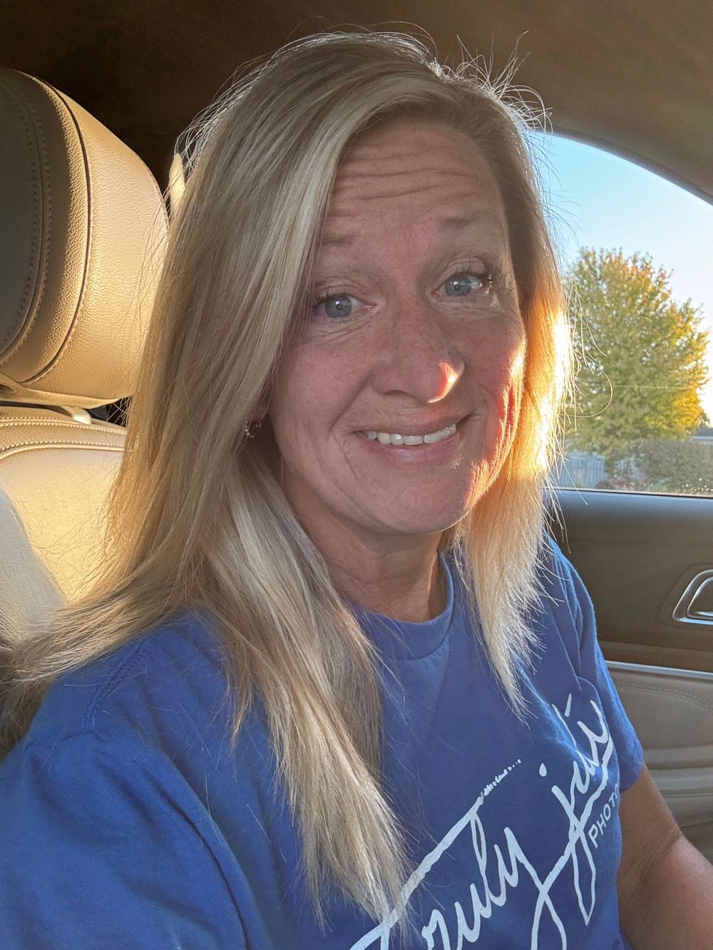 A selfie taken in a car showing a photographer wearing a blue Truly Juli Photography sweatshirt with blonde hair smiling at the camera.