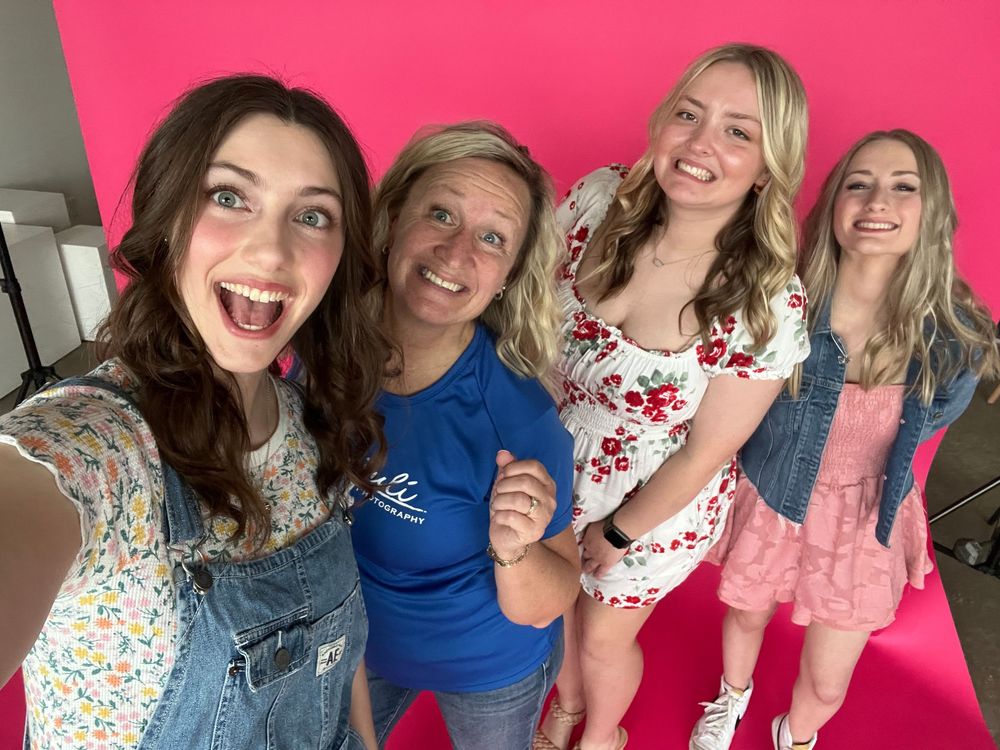 Photographer and her senior model team posing together for a selfie with excited expressions and bright smiles against a pink background.