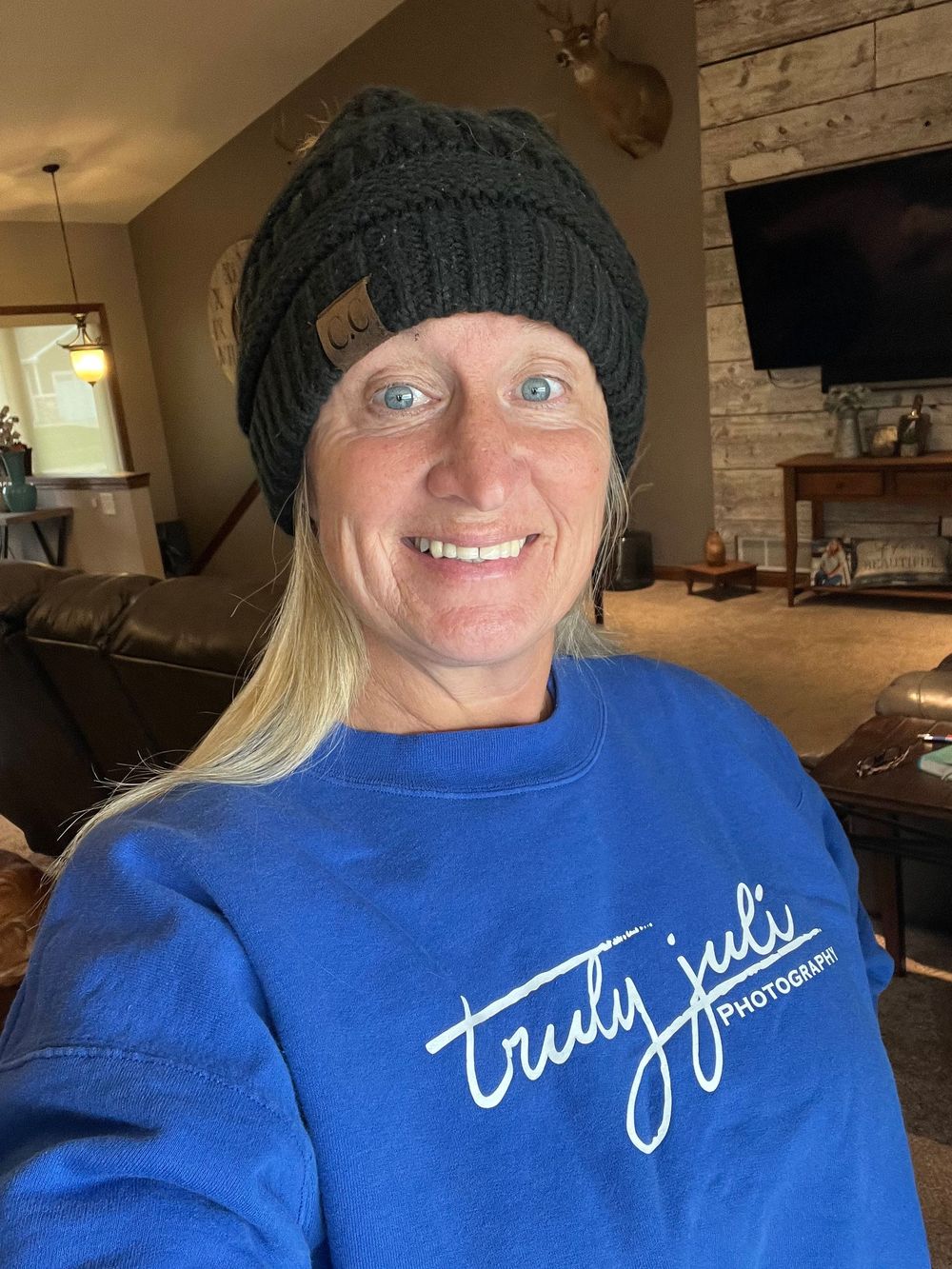 Someone wearing a blue sweatshirt with "Truly Juli" text and a dark knit beanie smiles at the camera in a cozy living room.