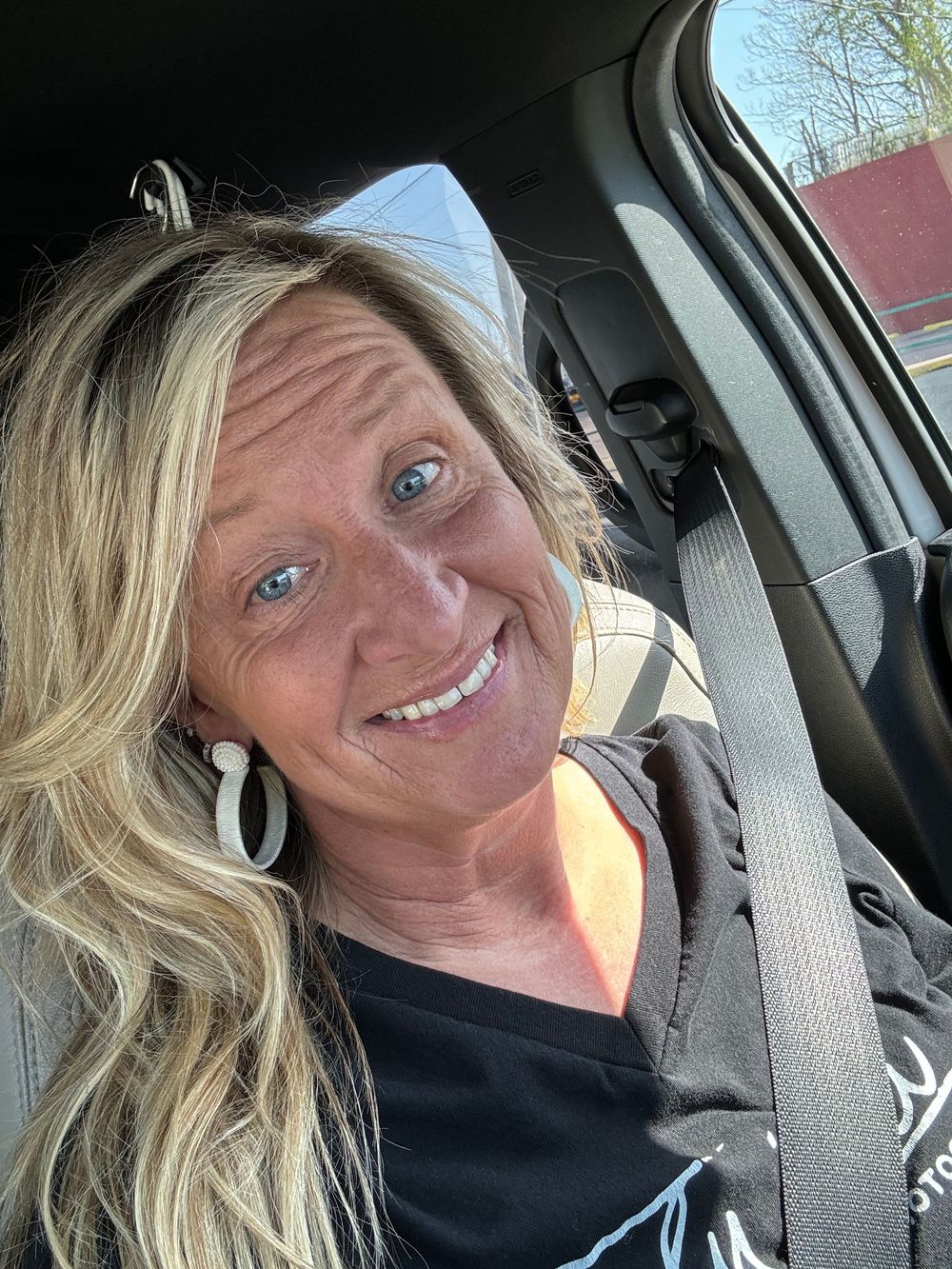 A selfie taken from inside a car showing a bright smile and blonde wavy hair against a sunny background.