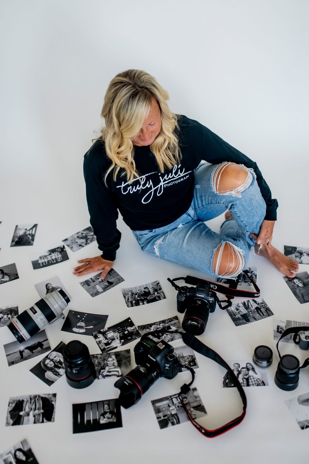 Photographer in black top and jeans arranges black and white photographs on white surface with vintage cameras nearby.