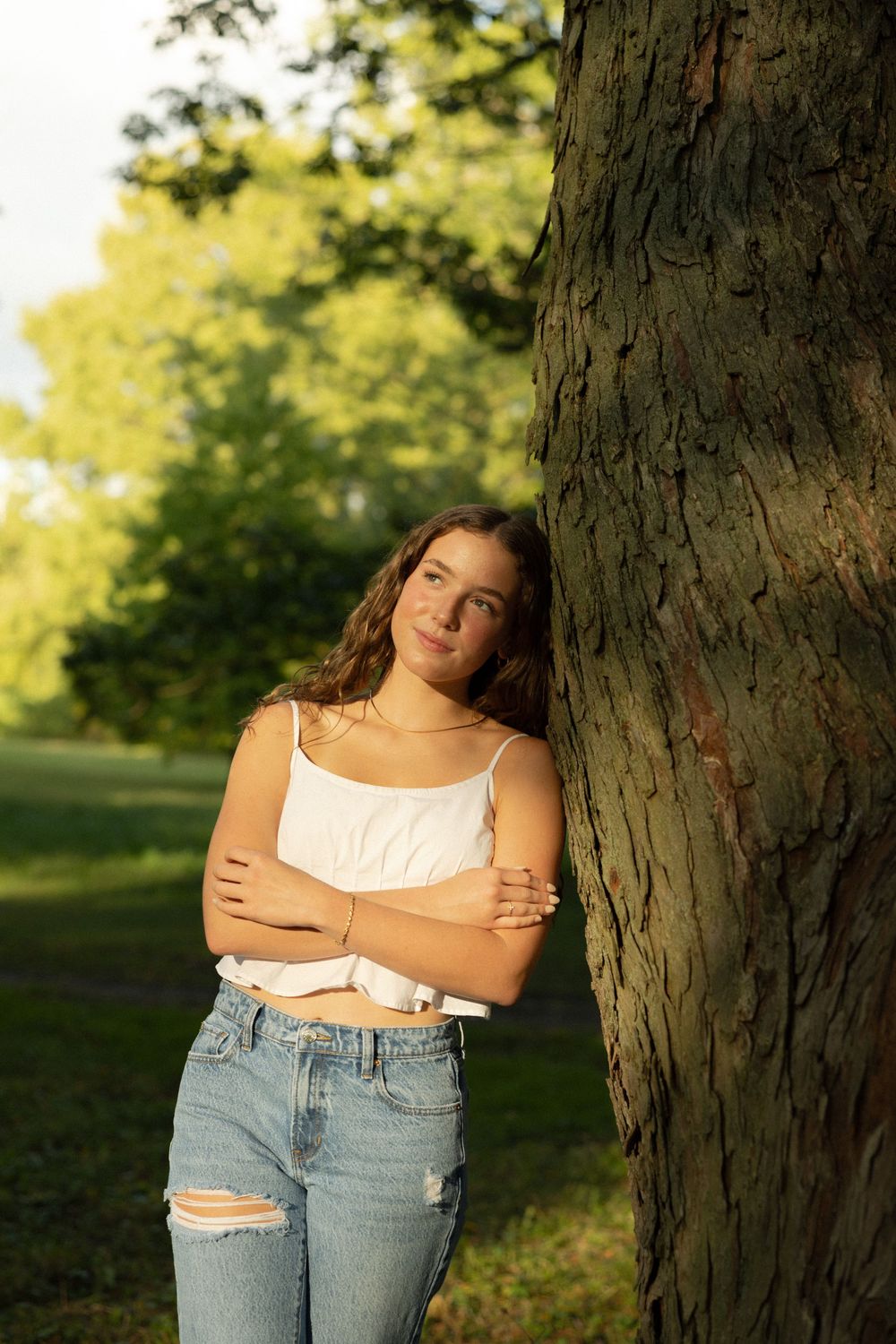 A person in a white tank top and ripped jeans poses against a large tree trunk in golden evening sunlight.