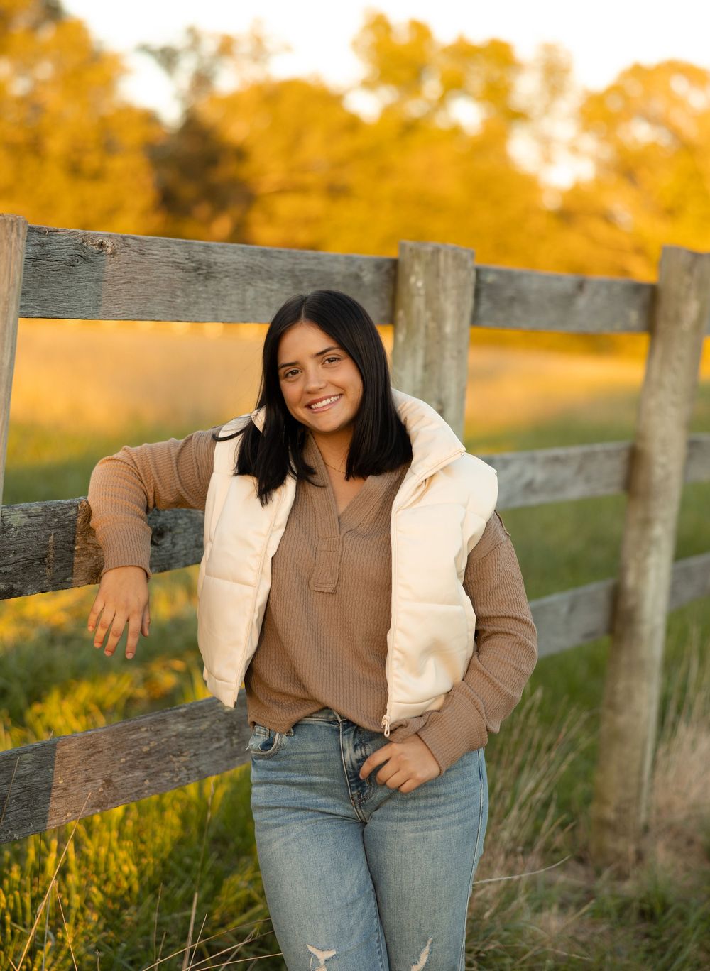 A person leans against a rustic wooden fence wearing a cream puffer vest and brown sweater on an autumn afternoon.
