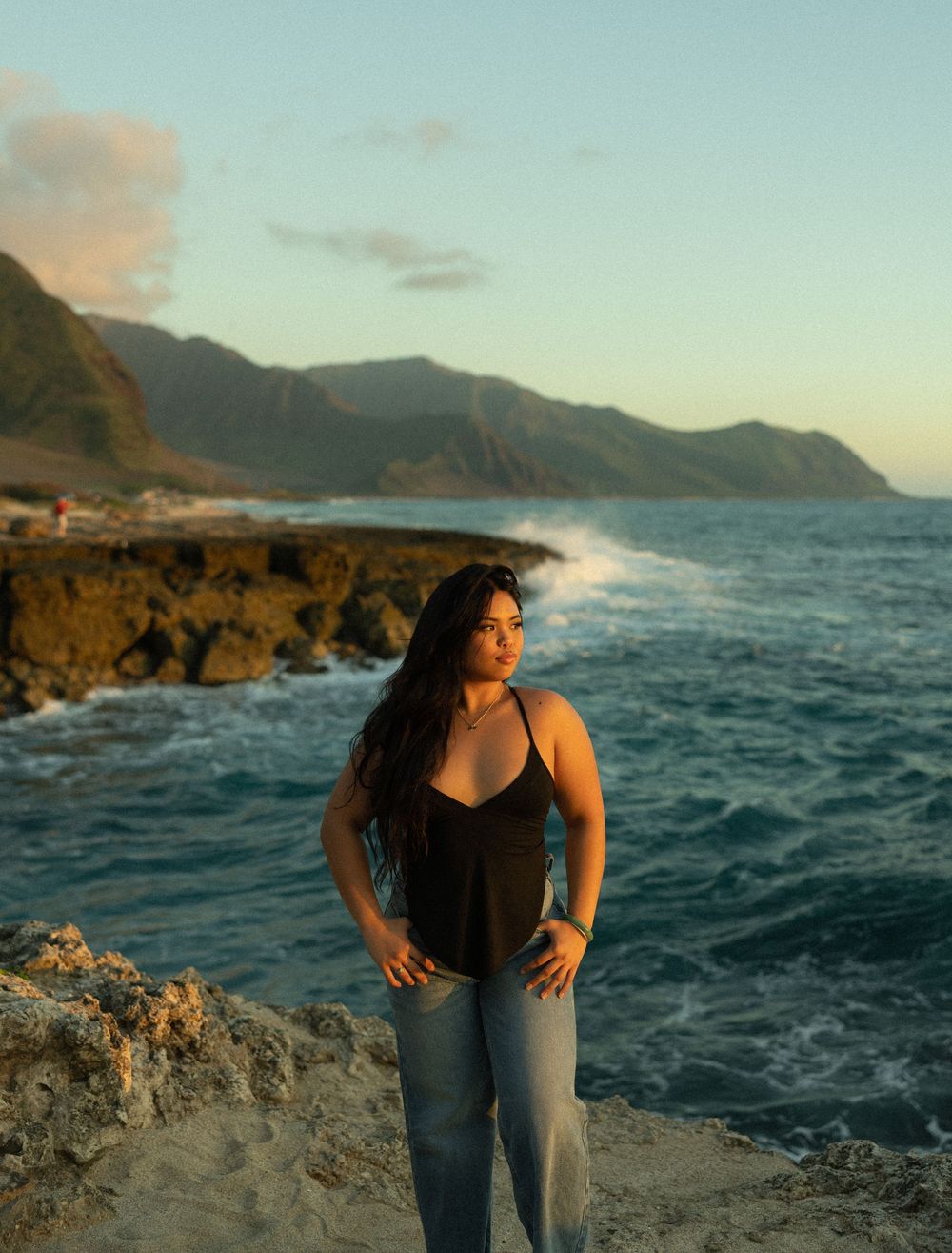 A person stands on rocky cliffs overlooking crashing waves against a mountainous coastline during golden hour.