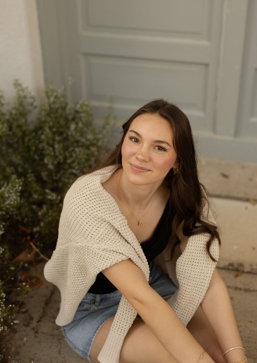 A young woman in a cream knit sweater and jeans sits casually outdoors next to decorative greenery.
