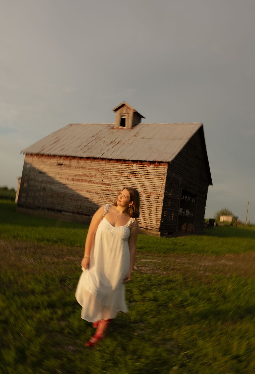 A person in a flowy white sundress and red boots stands near a weathered wooden barn at golden hour.