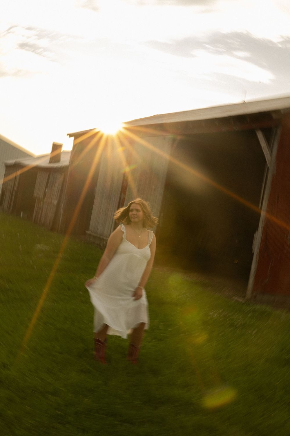 A figure in a flowing white dress dances in a grassy field at sunset with lens flare and barn buildings in background.