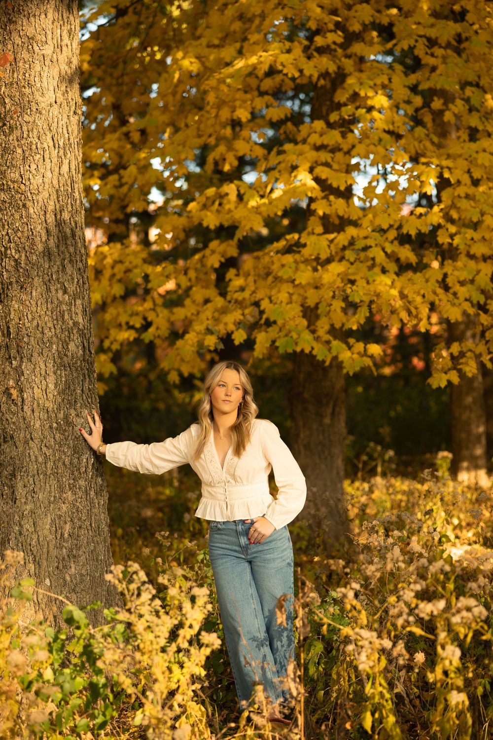 A person in a white blouse and jeans leans against a large tree trunk surrounded by golden autumn foliage.