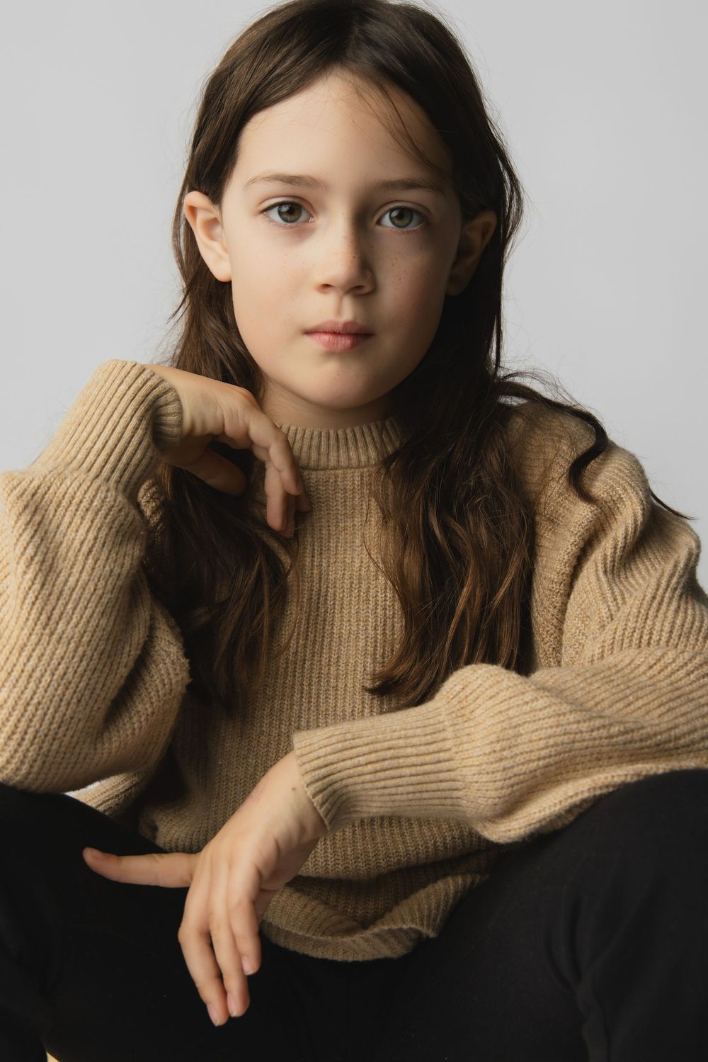 Young girl in a beige ribbed knit sweater sitting relaxed against a light gray background.