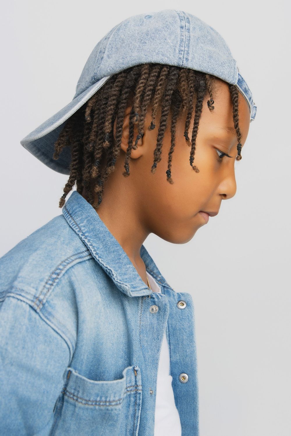 Young boy in denim jacket and light blue denim bucket hat with braided hair, looking down against gray background.