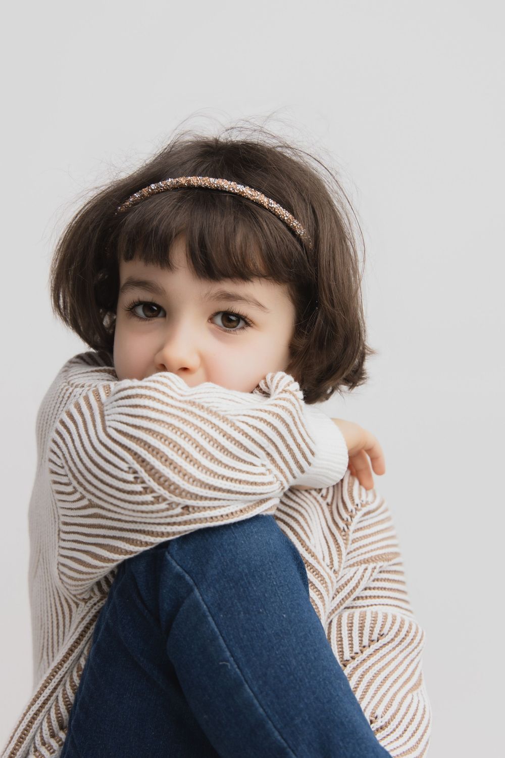 Young girl with dark bob hair and headband, hugging her knees in striped sweater and jeans, studio portrait.
