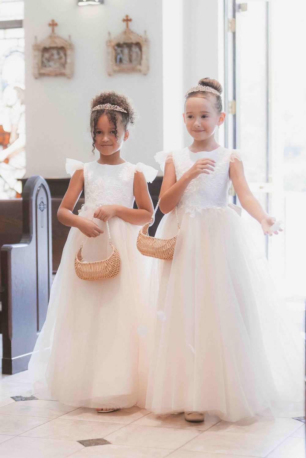 Two flower girls in white tulle dresses holding wicker baskets stand in a church aisle.