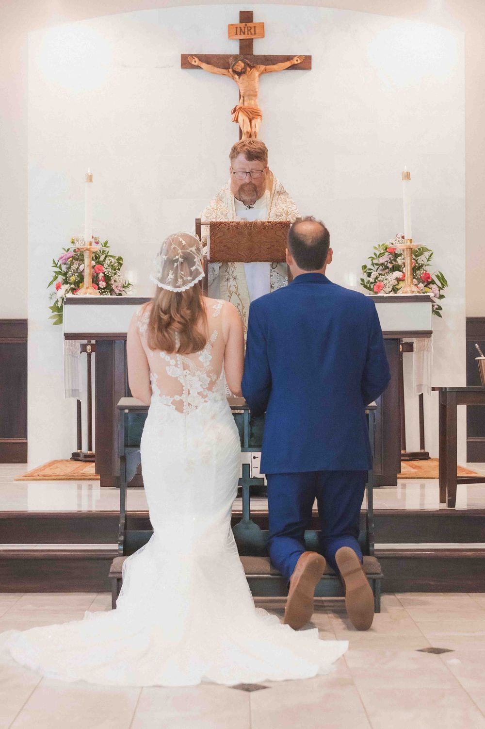 Bride and groom kneeling at church altar during Catholic wedding ceremony with priest and crucifix.