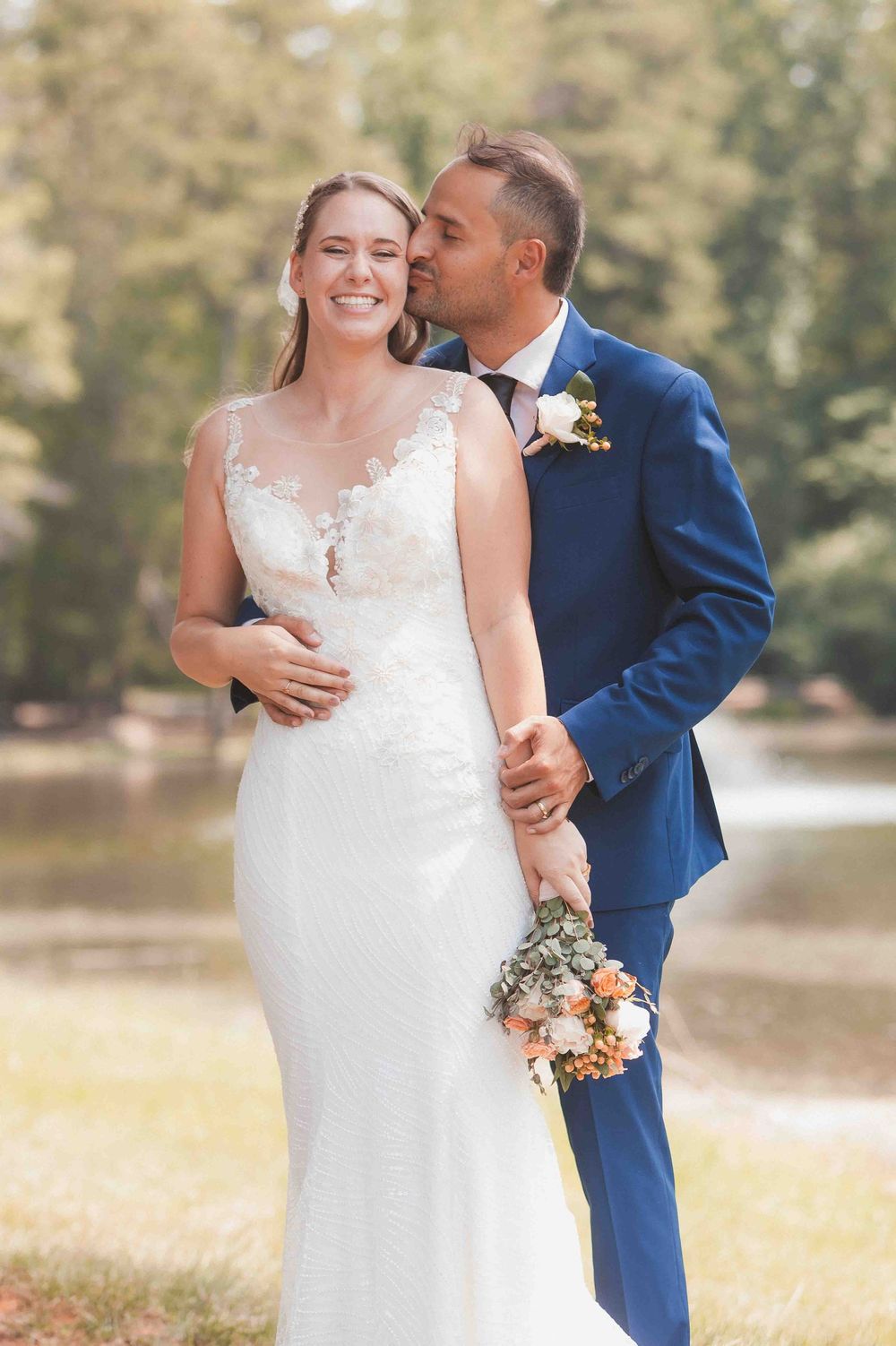 Bride in white lace gown holds floral bouquet as groom in navy suit kisses her cheek outdoors.