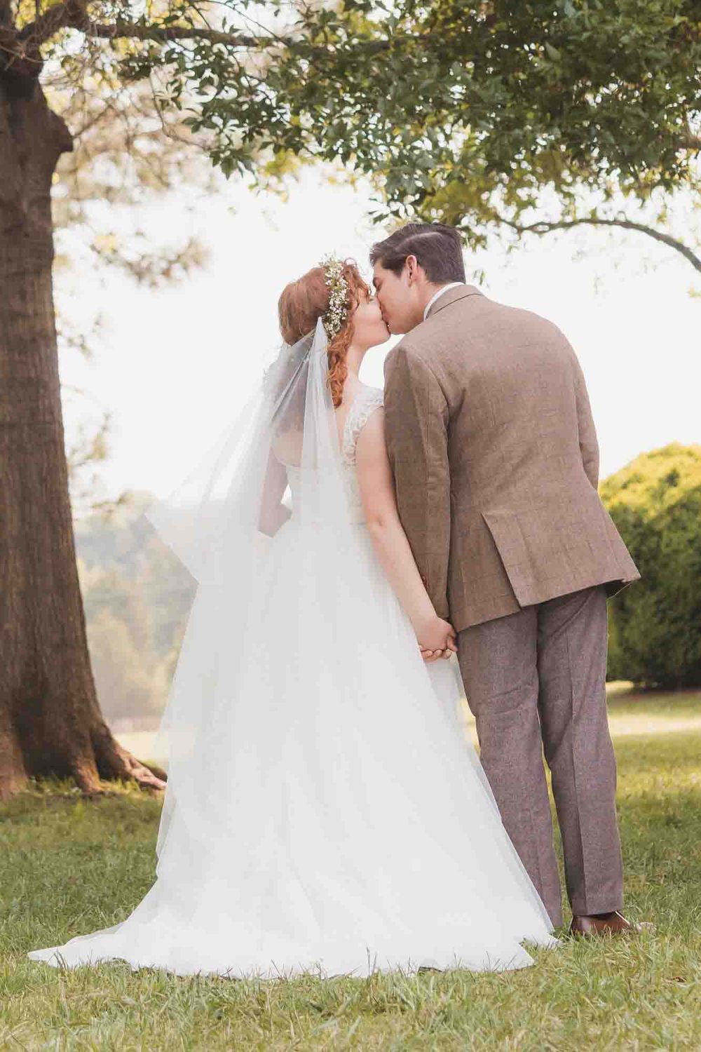 A bride and groom share a romantic kiss outdoors under a large tree, bride in white gown with veil.