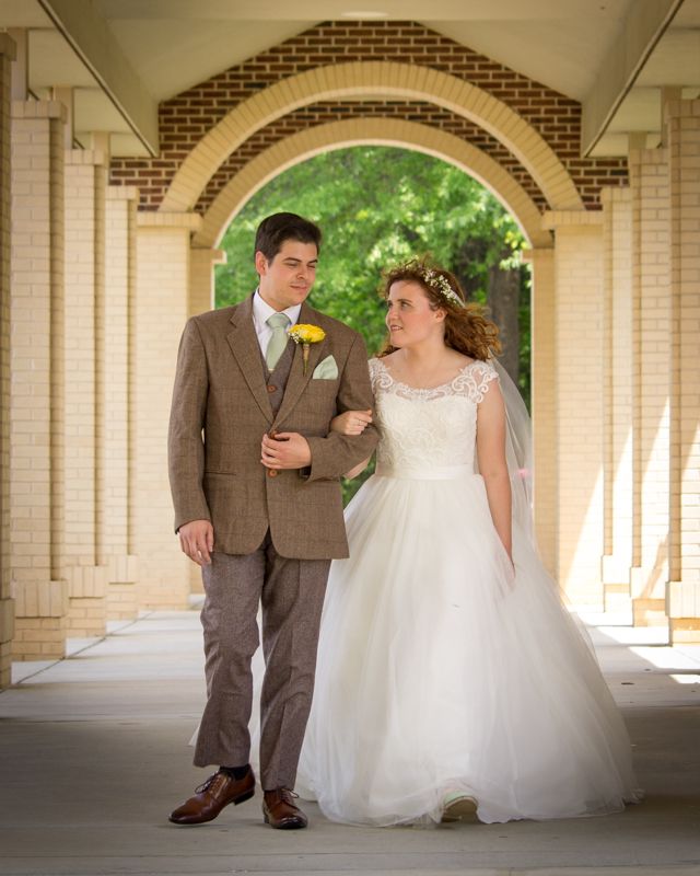 Bride and groom walking under brick arched colonnade, she in white ballgown, he in brown tweed suit with yellow boutonniere.