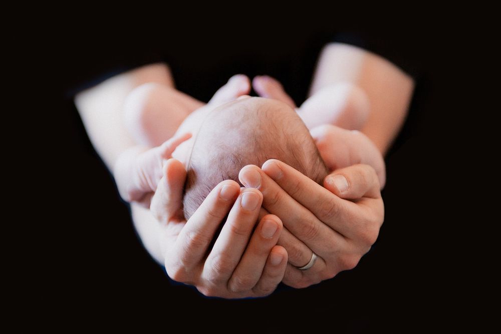Adult hands gently cradling a tiny newborn baby against a dark background.