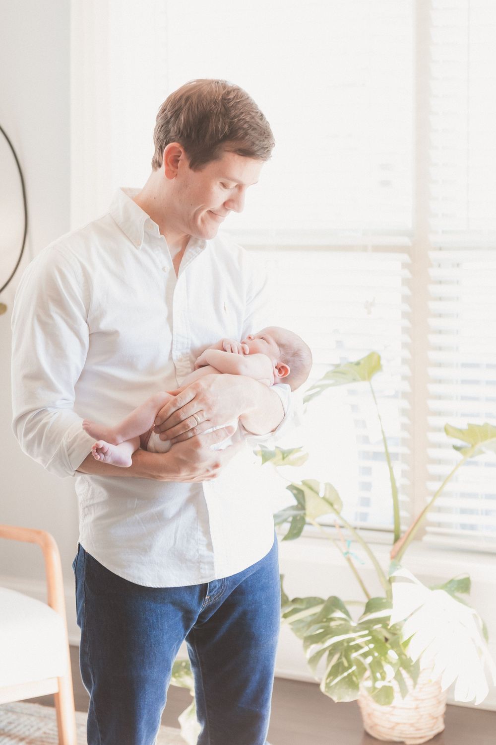 A father in a white shirt and jeans tenderly holds his newborn baby near a bright window with plants nearby.