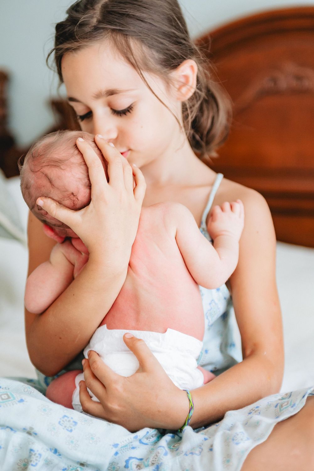 Young girl tenderly kissing a newborn baby while sitting on a bed, cradling the infant gently in her arms.